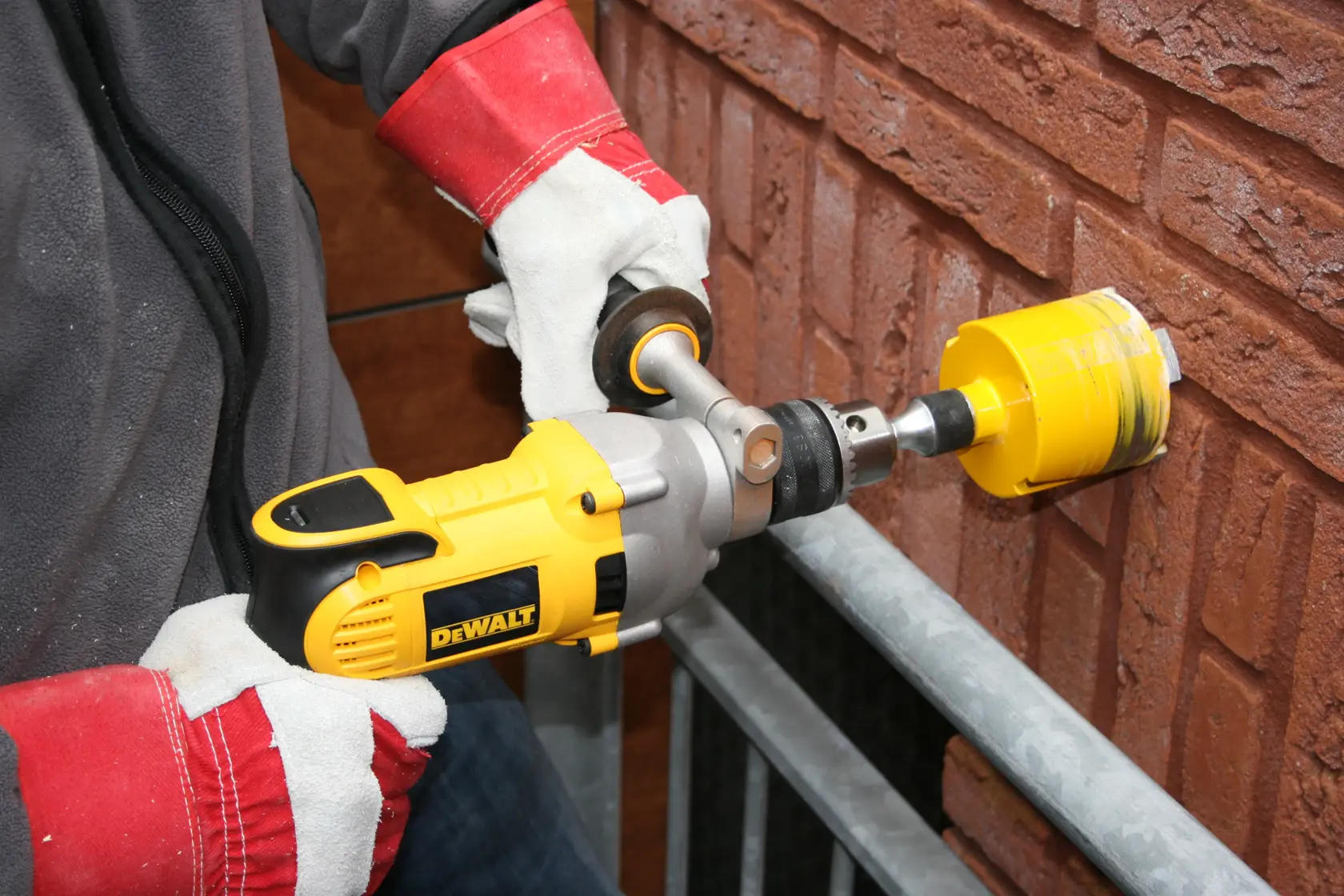 Eine Person mit Schutzhandschuhen verwendet einen gelben DEWALT-Elektrobohrer mit einem Lochsägenaufsatz, um in eine Ziegelwand zu bohren.