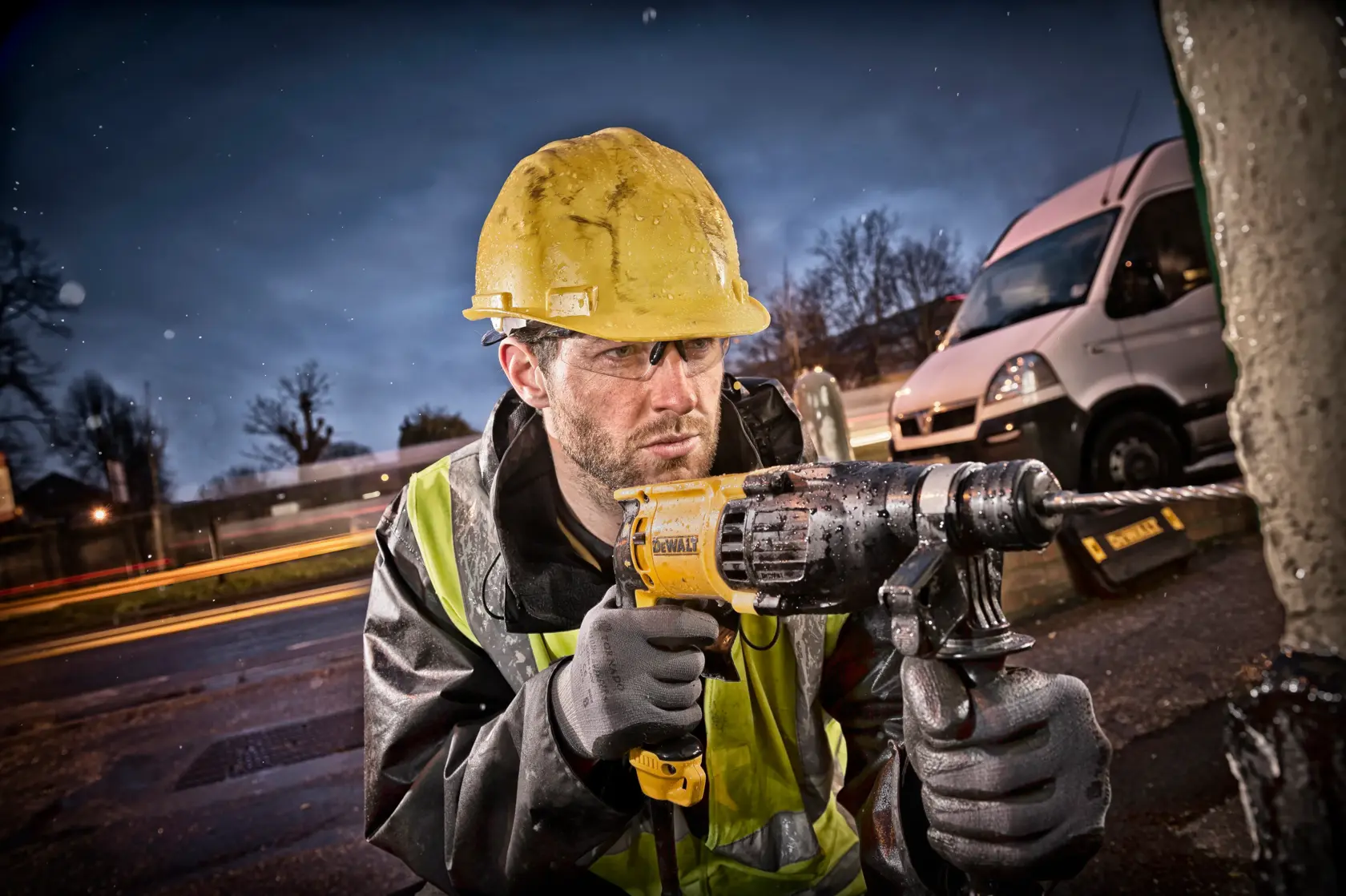 Ein Bauarbeiter mit gelbem Helm und reflektierender Jacke benutzt eine DEWALT-Schlagbohrmaschine im Freien, im Hintergrund sind ein weißer Lieferwagen und verschwommene Lichter zu sehen.