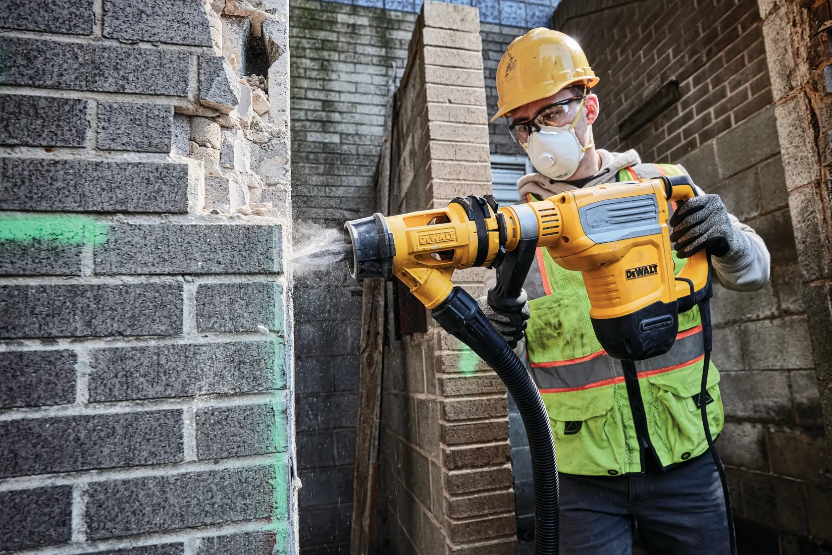 Ein Bauarbeiter mit Sicherheitshelm und Weste verwendet einen gelben DEWALT D25614K Abbruchhammer mit Staubabsaugung an einer Ziegelmauer.
