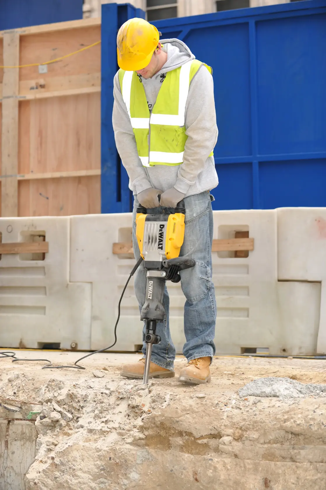 Une personne portant un casque jaune, des gants de sécurité et un gilet réfléchissant utilise un marteau-piqueur électrique DEWALT (modèle D25961K) pour casser du béton sur un chantier extérieur.