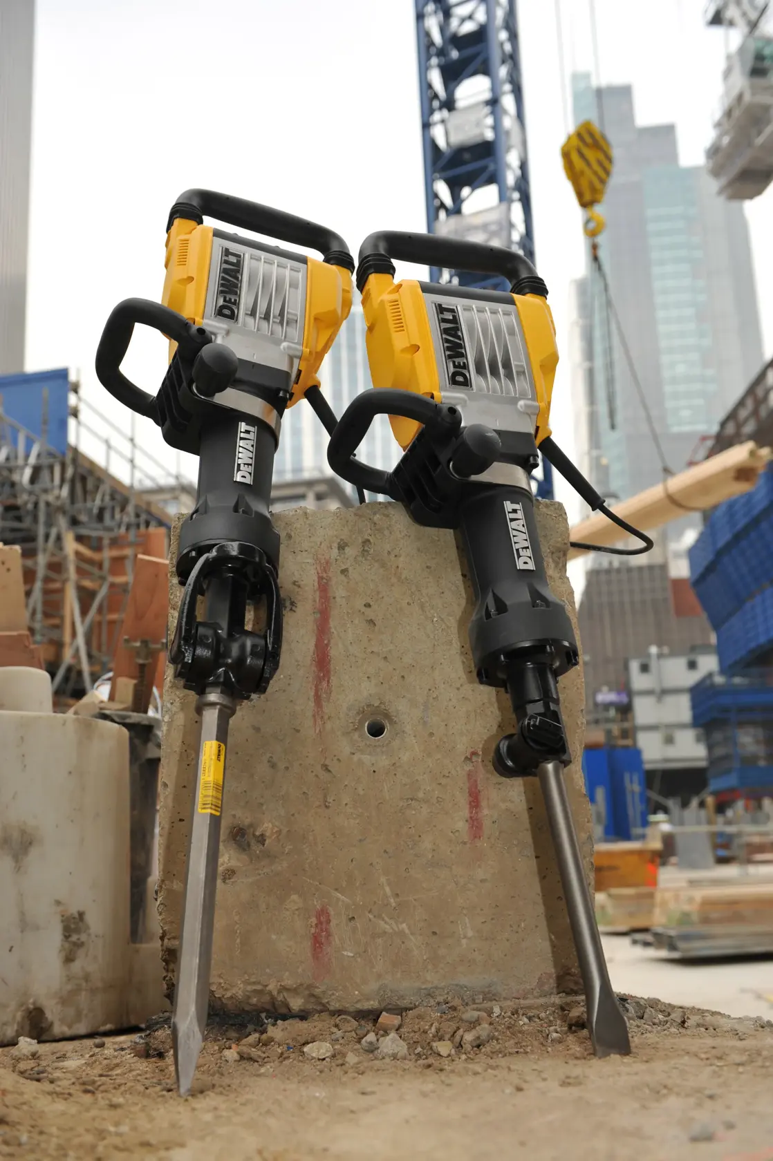 Deux marteaux-piqueurs électriques DEWALT sont posés verticalement sur un bloc de béton sur un chantier, avec des immeubles et des grues en arrière-plan.
