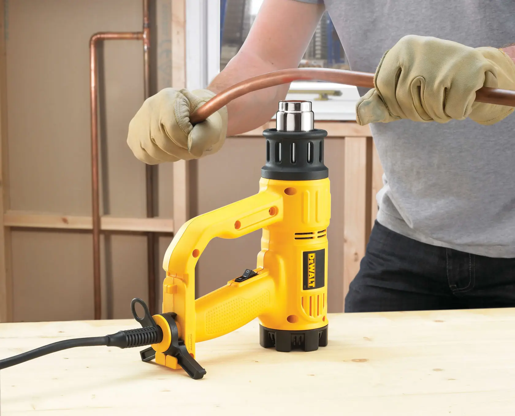 Eine Person mit Handschuhen verwendet eine DEWALT-Heißluftpistole, um ein Kupferrohr auf einer Holzarbeitsfläche zu biegen.