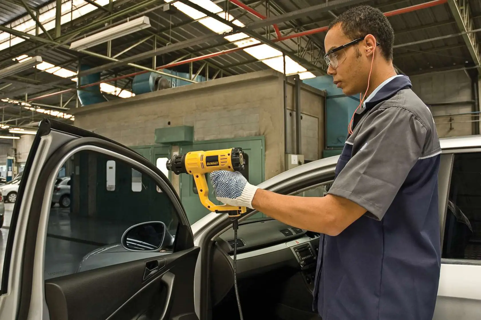 Eine Person mit Handschuhen verwendet eine gelbe DEWALT Heißluftpistole (Modell D26411) im Innenbereich einer Autotür in einer Werkstatt. Das Gesicht der Person ist aus Datenschutzgründen unscharf.