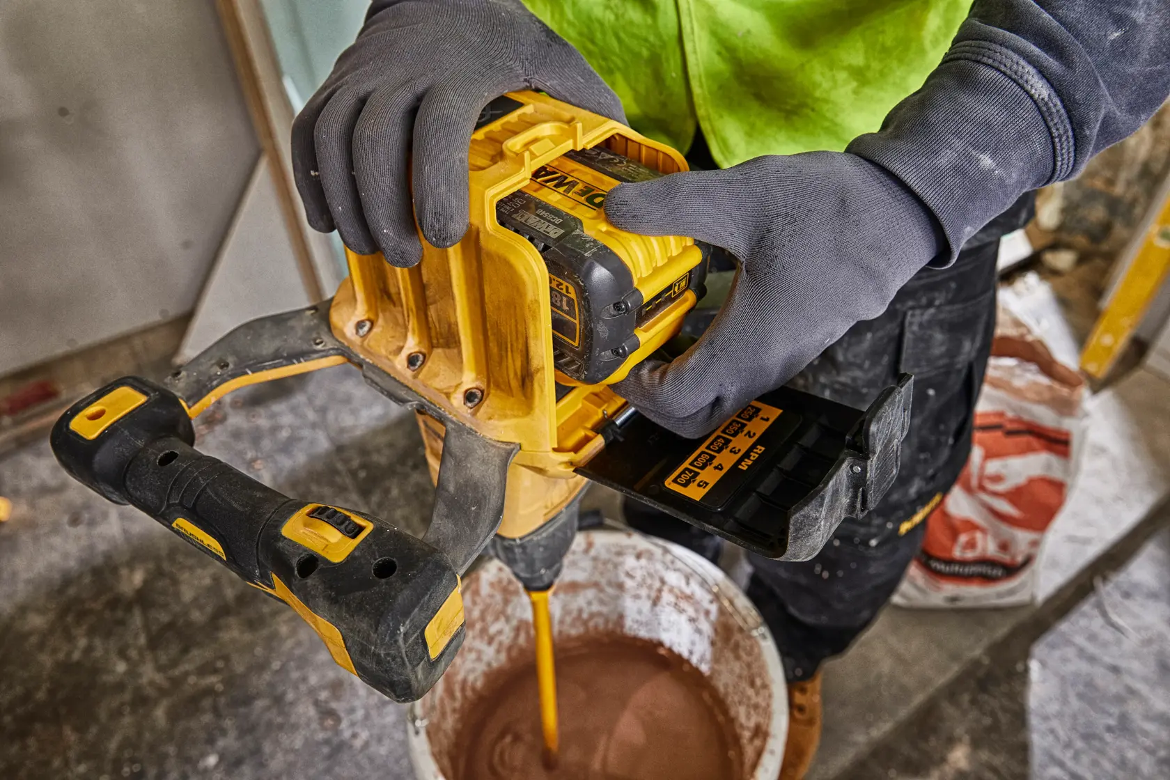 Una persona con i guanti utilizza un miscelatore cordless giallo DEWALT con una batteria DEWALT DCB548 per mescolare materiale in un secchio su un cantiere.