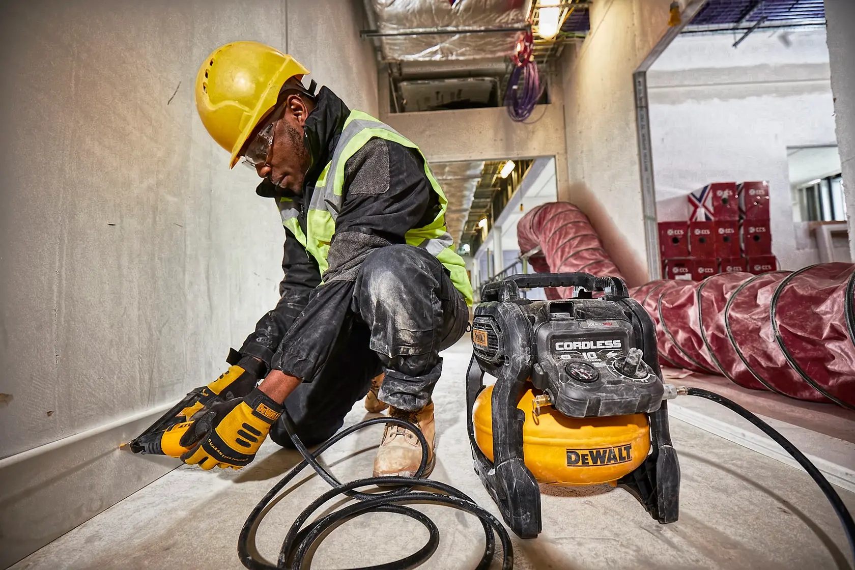 Un ouvrier du bâtiment portant un casque de sécurité jaune et un gilet de sécurité est agenouillé sur un chantier, utilisant un outil électrique relié à un compresseur d'air sans fil DEWALT modèle DCC1054N. Le compresseur est posé au sol, et l'ouvrier travaille le long d'un mur. L'environnement montre des équipements et matériaux industriels.