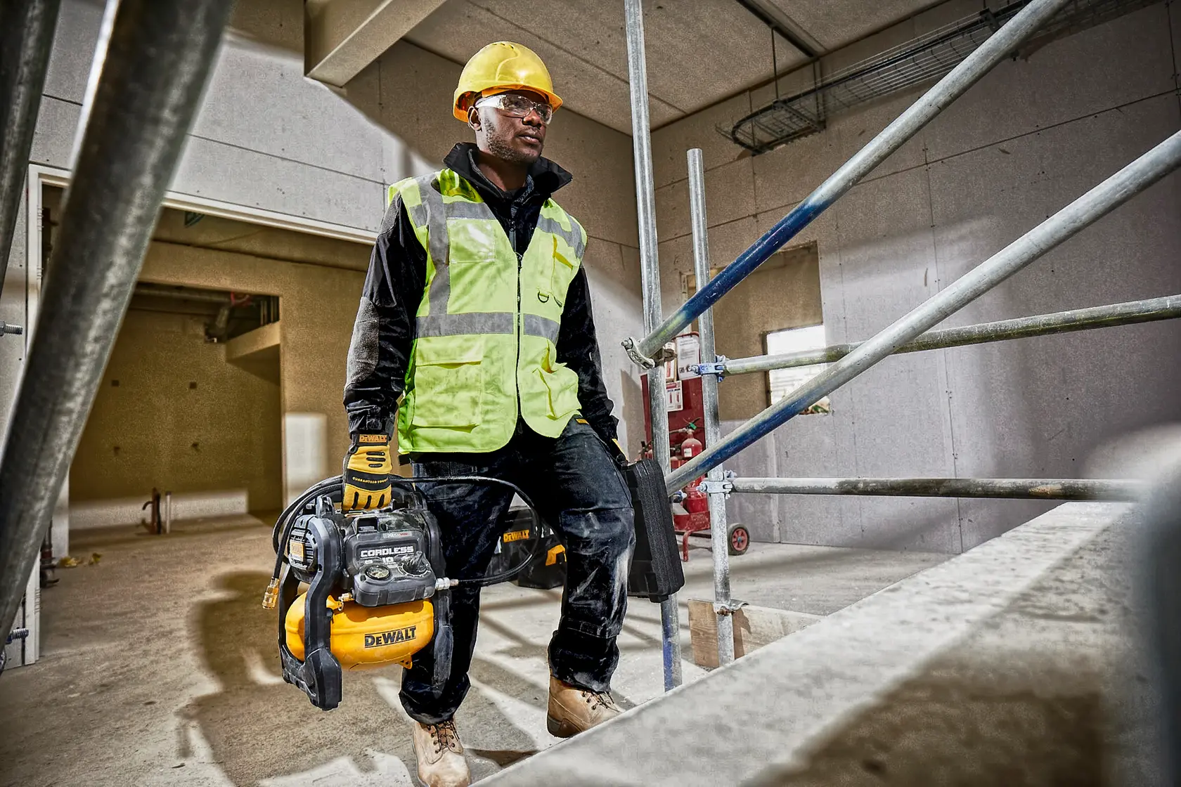 Un ouvrier du bâtiment portant un casque de chantier jaune, un gilet haute visibilité, des gants et des chaussures de sécurité porte un compresseur d'air portable DEWALT (modèle DCC1054N) sur un chantier. Le visage de l'ouvrier est flouté pour préserver sa vie privée. L'environnement est en construction avec des échafaudages et des murs non finis.