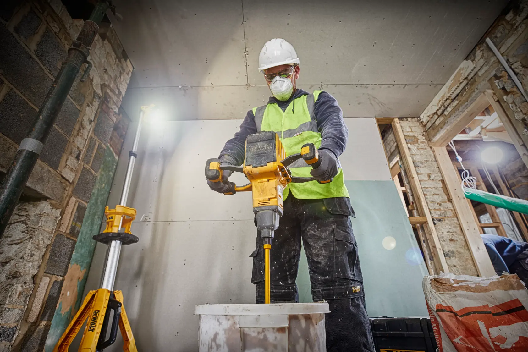 Un trabajador de la construcción con casco y chaleco de alta visibilidad usa un mezclador DEWALT DCD240X2 para mezclar material en un cubo, dentro de un edificio sin terminar.