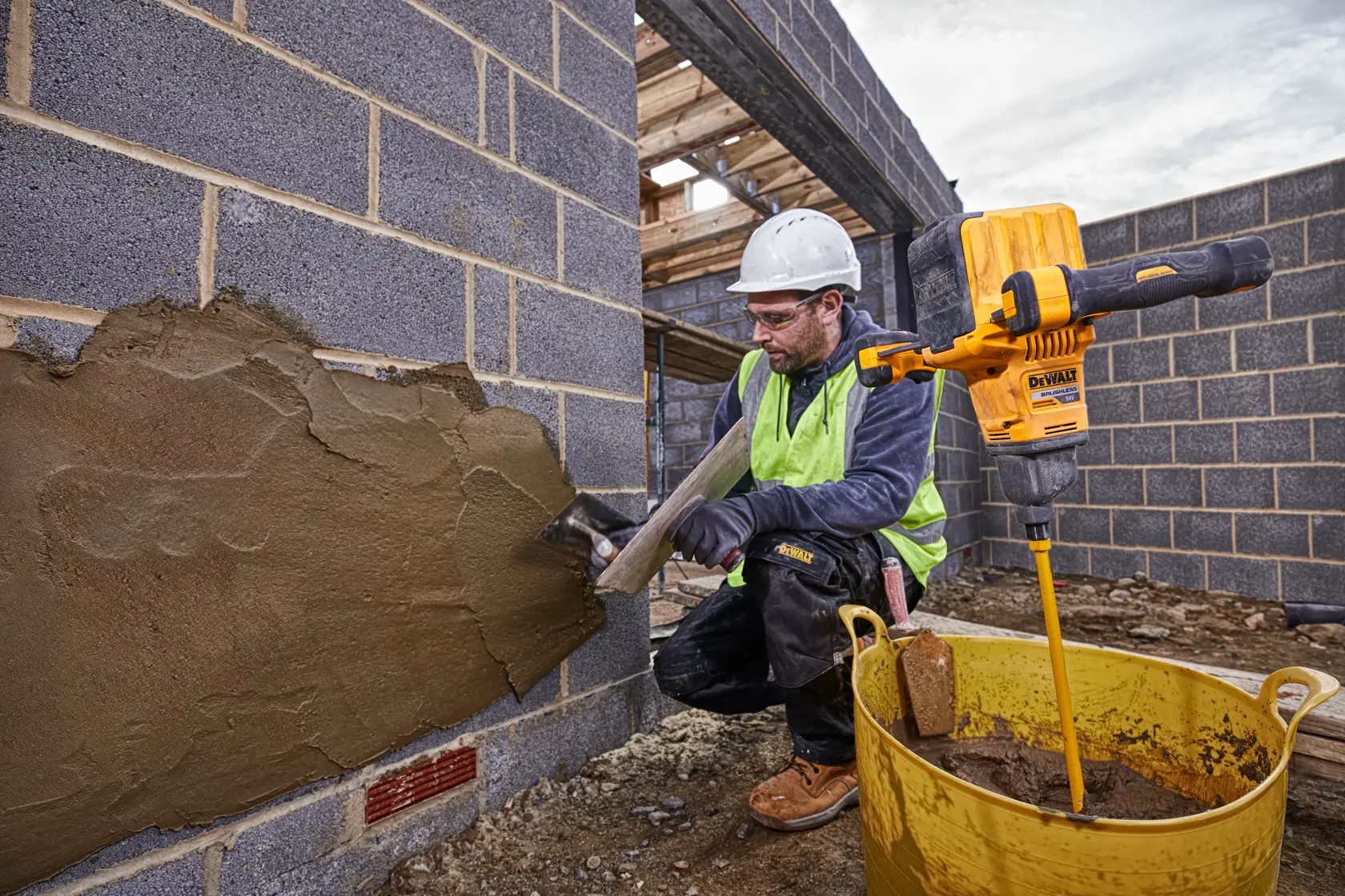 Un trabajador de la construcción con casco de seguridad y chaleco reflectante aplica yeso a una pared de ladrillos en una obra. Junto al trabajador hay un taladro mezclador amarillo DEWALT (modelo DCD240X2) colocado en un gran recipiente amarillo lleno de mezcla de yeso.
