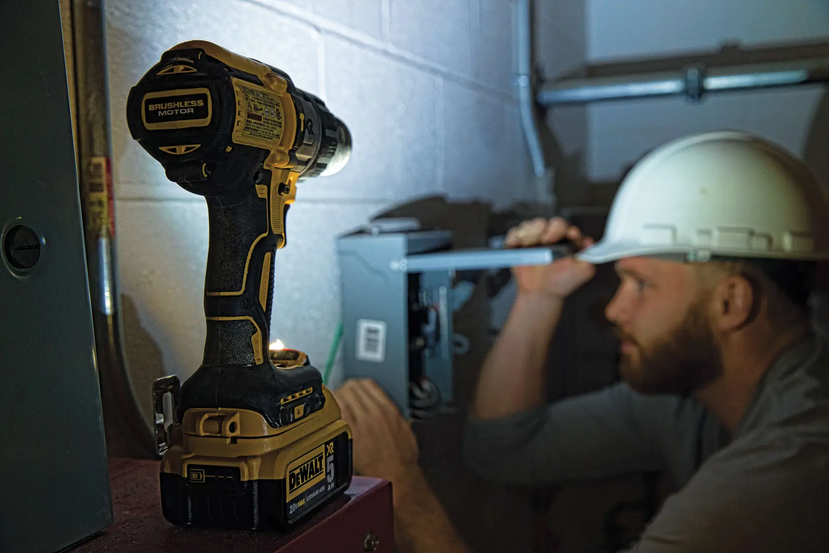 Una persona con giacca ad alta visibilità e casco da cantiere sta utilizzando un trapano avvitatore a batteria DEWALT (modello DCD996P2) per forare un muro in cemento o muratura in un ambiente industriale o edile. Il prodotto e la presa sull’utensile sono chiaramente visibili.