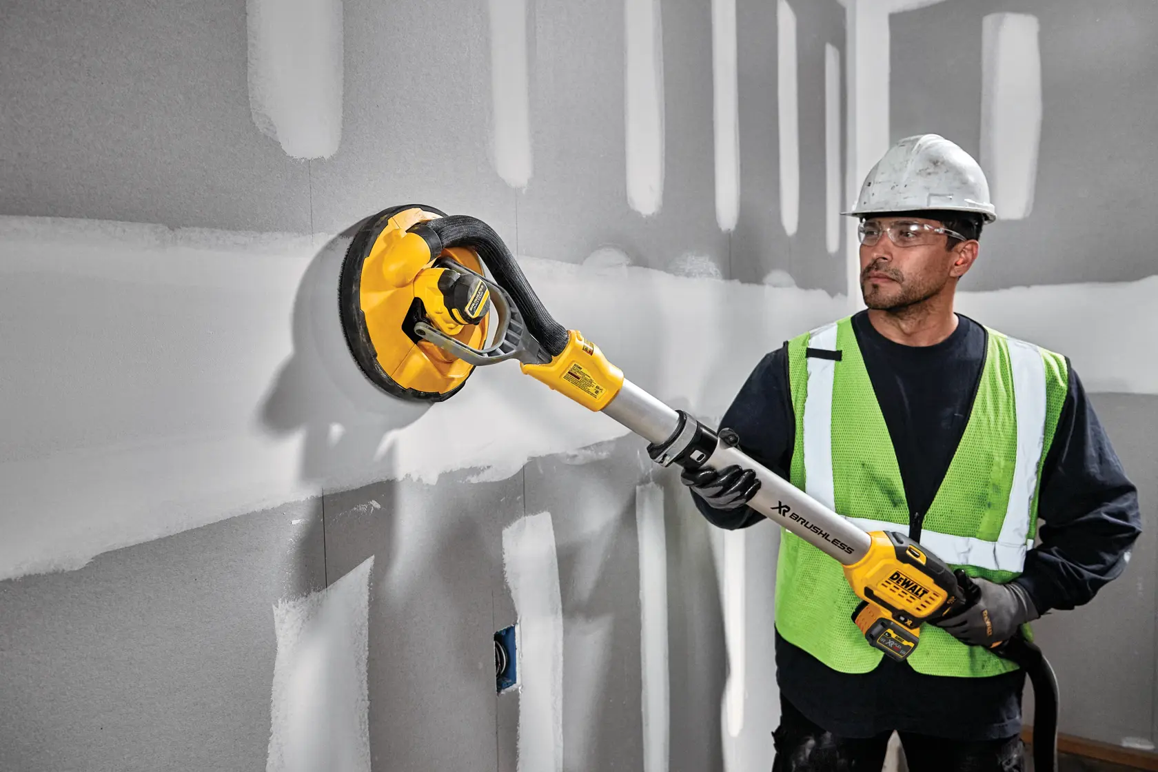 Una persona con casco e gilet di sicurezza utilizza una levigatrice per cartongesso senza fili DEWALT DCE800T2 su una parete con giunti di intonaco non finiti.