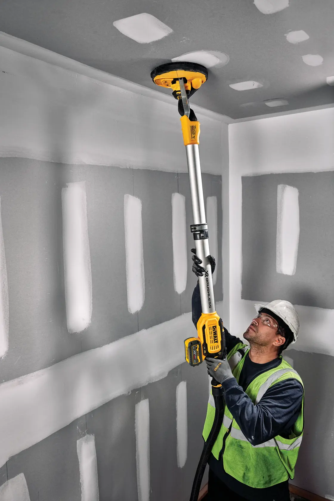 Un operatore con gilet di sicurezza e casco utilizza una levigatrice per cartongesso a batteria DEWALT con asta di prolunga per carteggiare il soffitto in una stanza con cartongesso non finito.