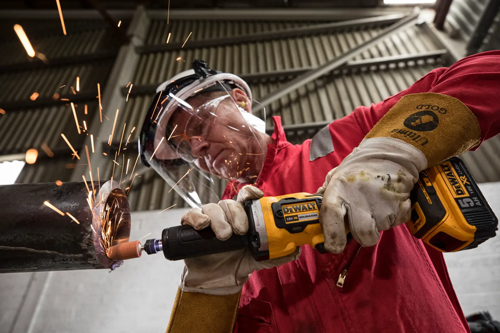 Una persona indossa guanti protettivi e dispositivi di sicurezza mentre utilizza una smerigliatrice senza fili DEWALT DCG426N su un tubo metallico, generando scintille in un ambiente industriale.