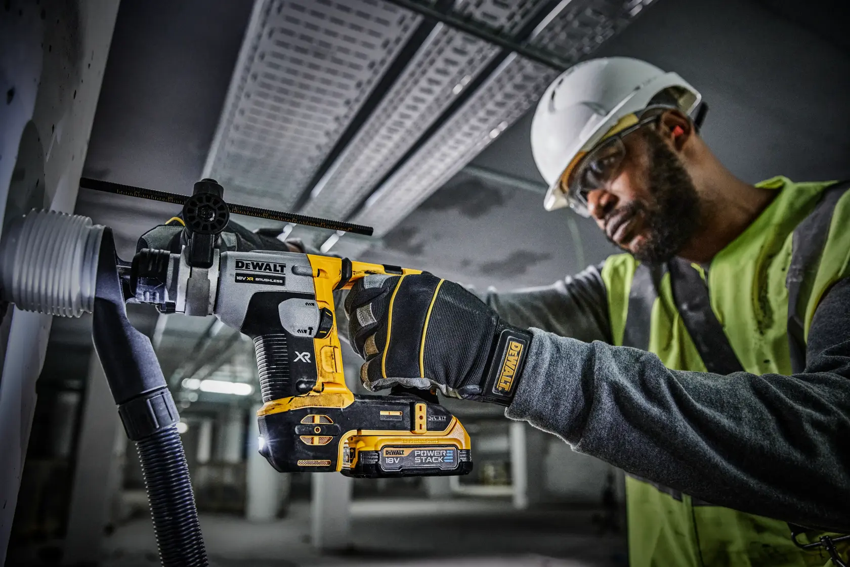 Una persona con casco di sicurezza e giubbotto ad alta visibilità sta usando un martello rotativo a batteria DEWALT per forare una parete, con un sistema di aspirazione della polvere collegato. Il martello presenta le scritte XR e Power Stack.