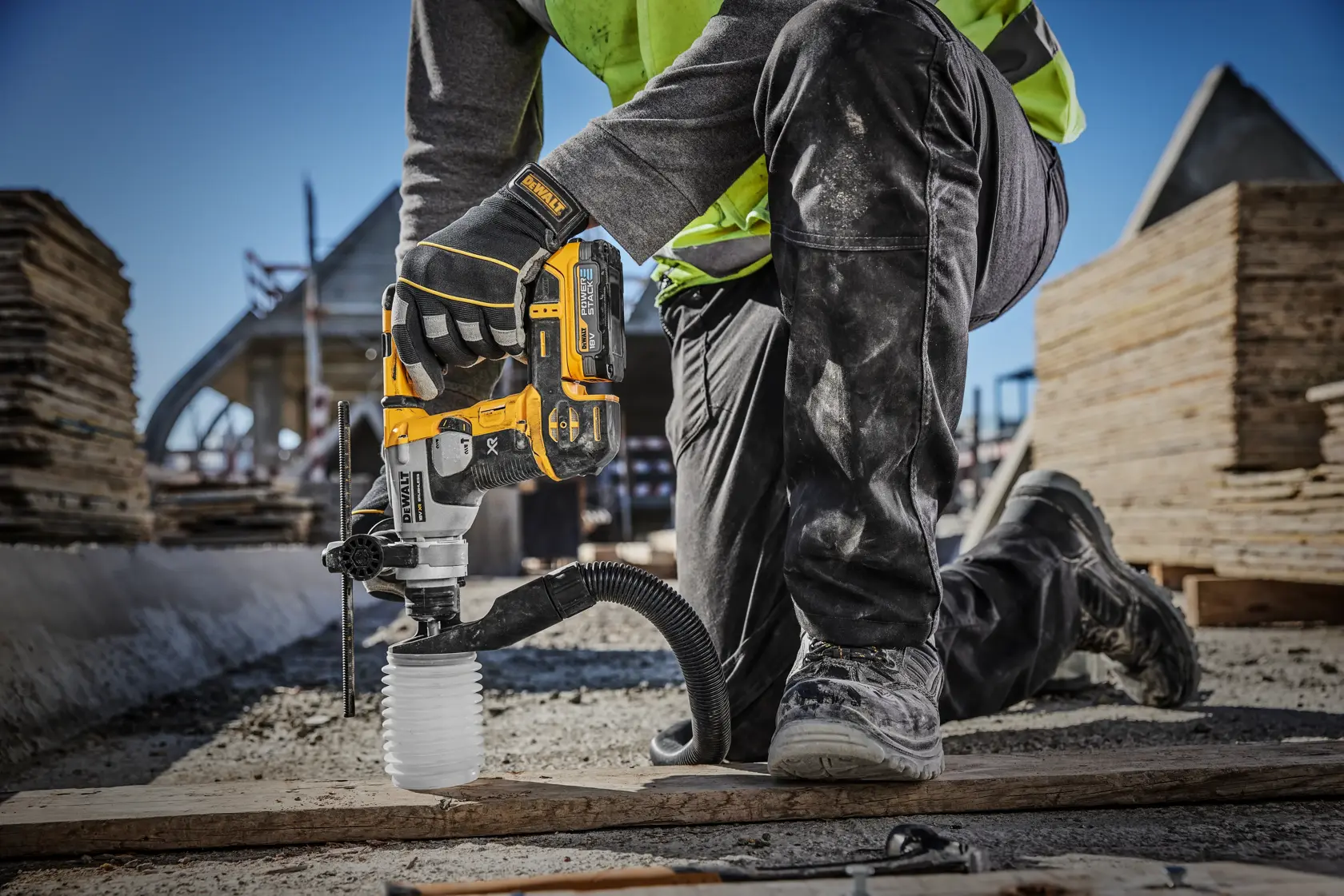 Un operaio edile utilizza un martello perforatore a batteria DEWALT (modello DCH172E2T) con aspirazione della polvere in un cantiere, forando il terreno in ginocchio.