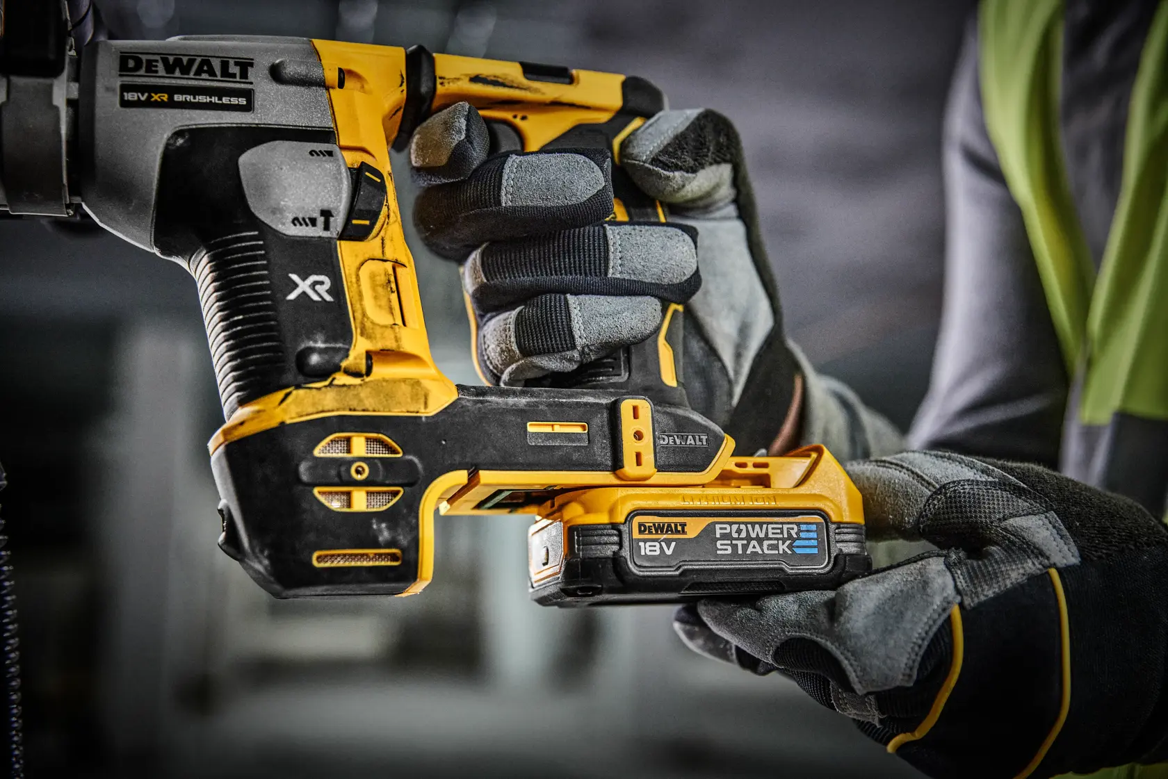 Primo piano di una persona con guanti da lavoro che tiene un utensile elettrico DEWALT 18V XR brushless con batteria Power Stack in un ambiente di officina o cantiere.