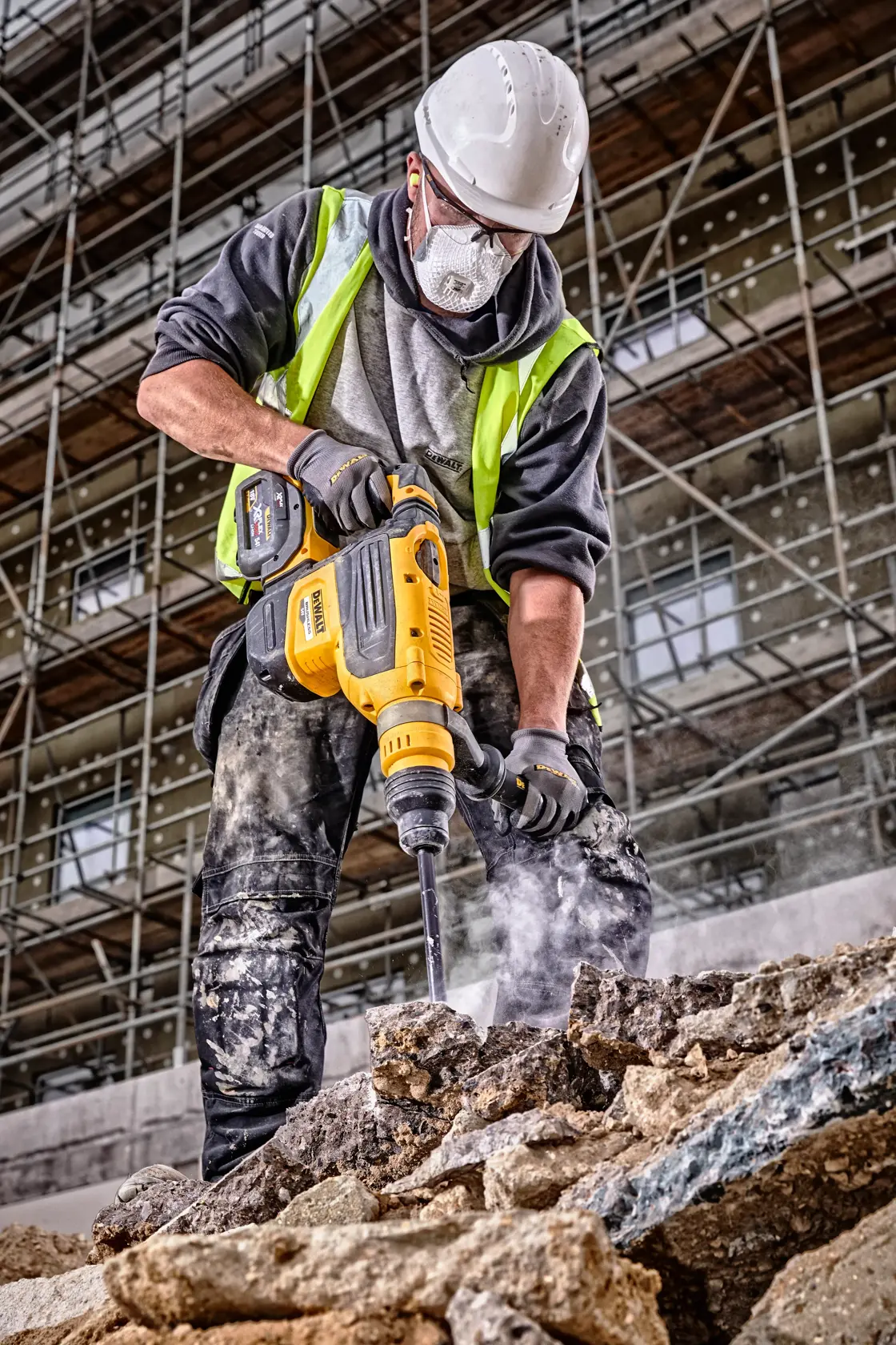Un operaio edile con casco e giubbotto riflettente utilizza un martello rotativo DEWALT DCH733N per rompere detriti di cemento in un cantiere, con impalcature visibili sullo sfondo.