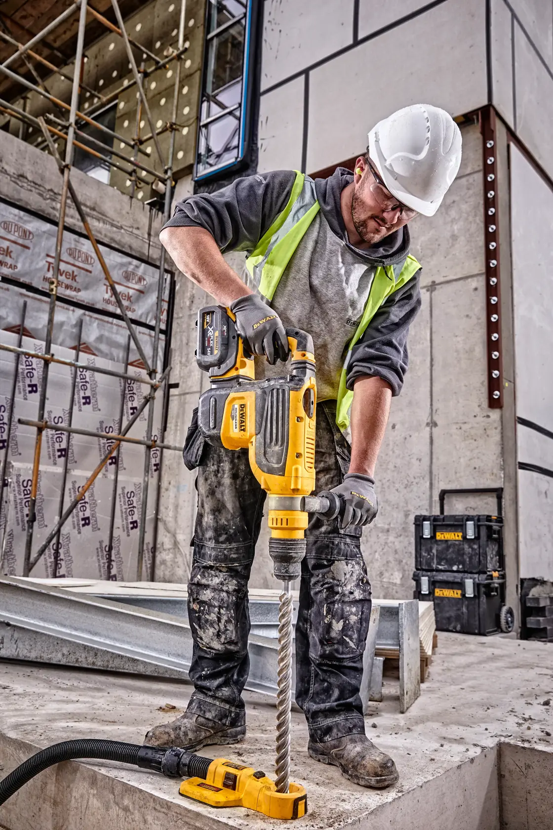 Un operaio edile con elmetto e gilet di sicurezza utilizza un martello rotante DEWALT DCH733N su una superficie di cemento in un cantiere. In background si vedono scatole degli attrezzi DEWALT e impalcature.
