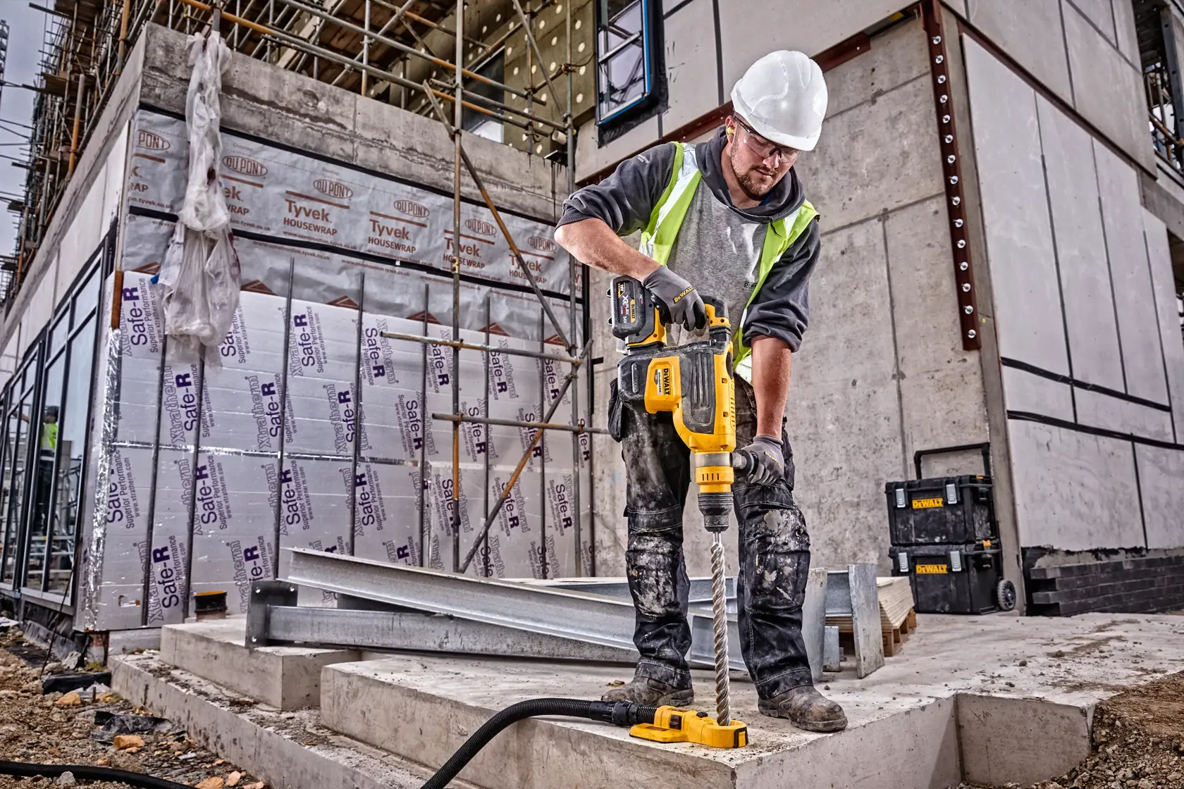 Un operaio edile con dispositivi di sicurezza utilizza un trapano rotativo giallo DEWALT (SKU: DCH733N) su una superficie in cemento in un cantiere.