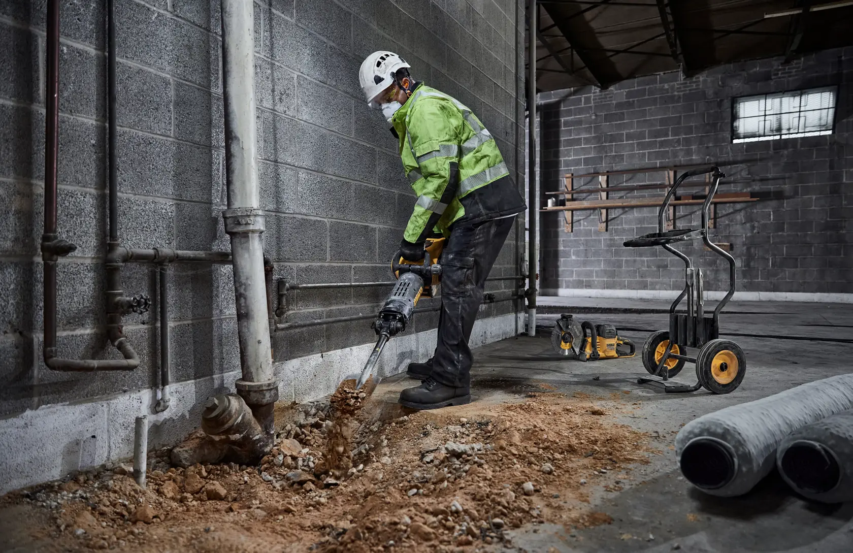 Un operaio edile con dispositivi di sicurezza utilizza un martello demolitore elettrico DEWALT per rompere il cemento in un cantiere. L’attrezzo è collegato a un sistema di aspirazione ed è presente attrezzatura da costruzione sullo sfondo.