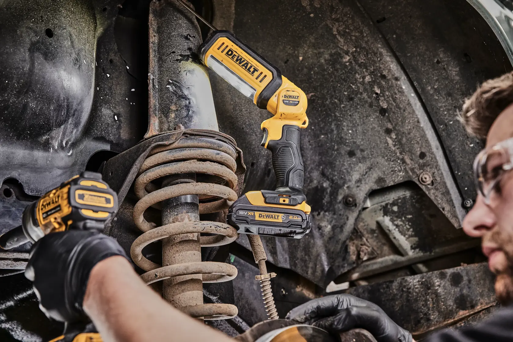 Una persona è inginocchiata e lavora su un'unità elettrica aperta usando degli attrezzi. Sul pavimento c'è una lampada da lavoro LED portatile DEWALT DCL050 che illumina l'area di lavoro.