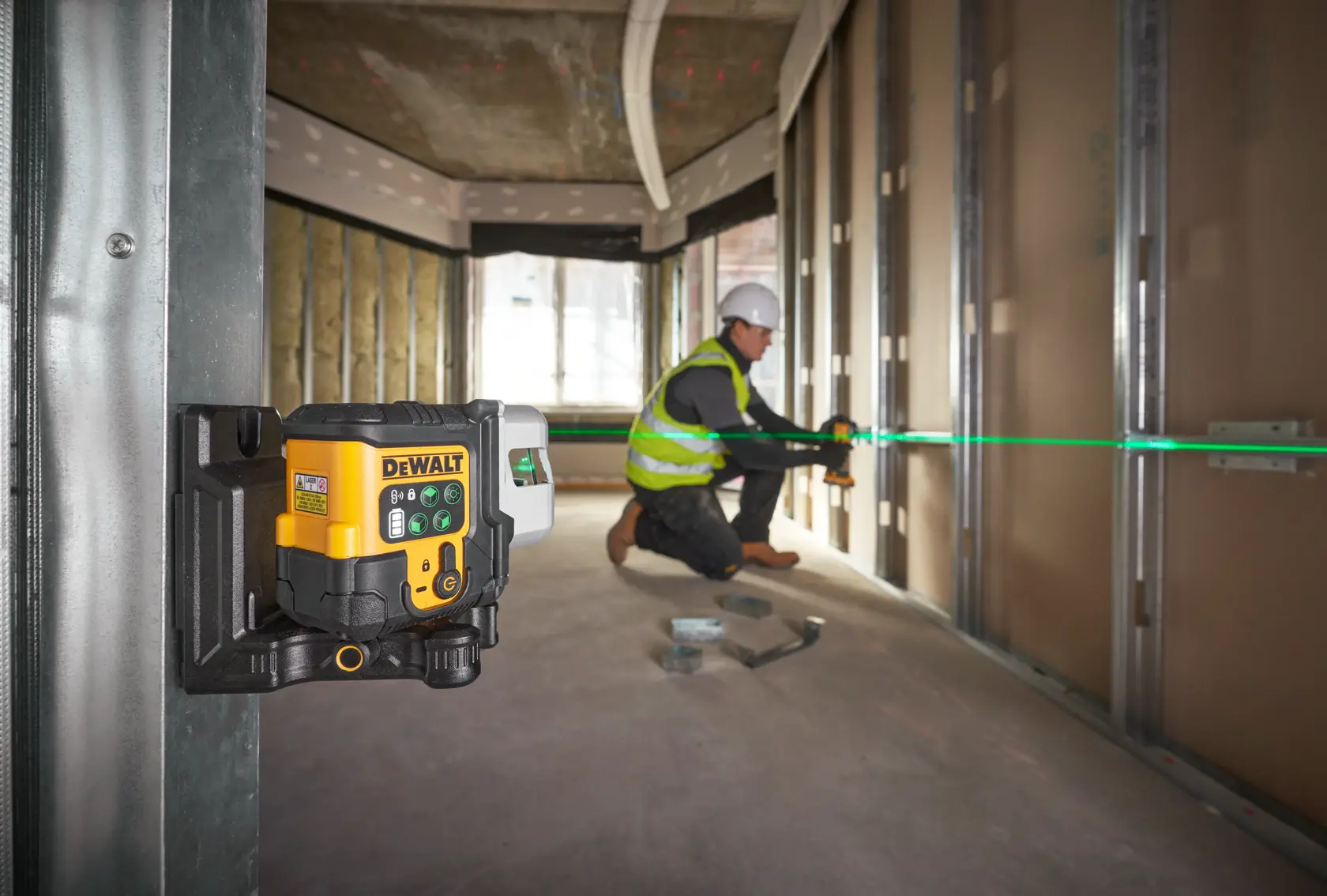 A DEWALT laser level device (SKU: DCLE14301GB) mounted on a metal frame inside a building under construction. The laser projects a green line across the room, while a person in safety gear works with tools along the line.