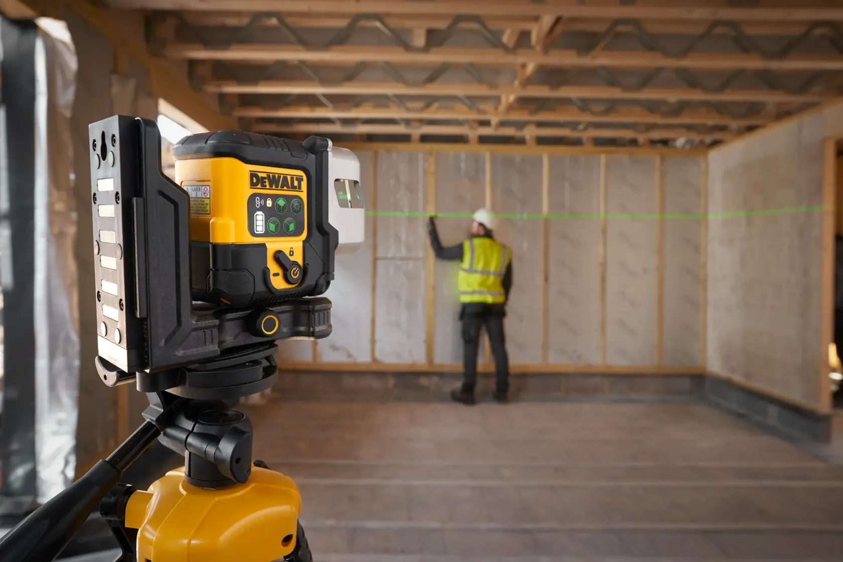 A DEWALT laser level device mounted on a tripod is in use at a construction site, projecting a green horizontal laser line. A person wearing a safety vest and helmet is working near a wall in the background.