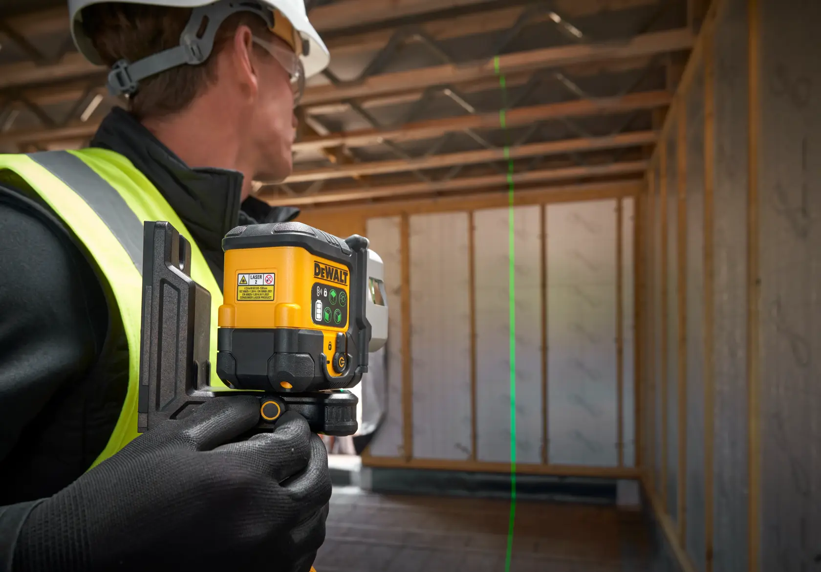 A person wearing safety gear uses a DEWALT laser level tool inside a wooden-frame construction site. The tool is projecting a green laser line onto the wall.