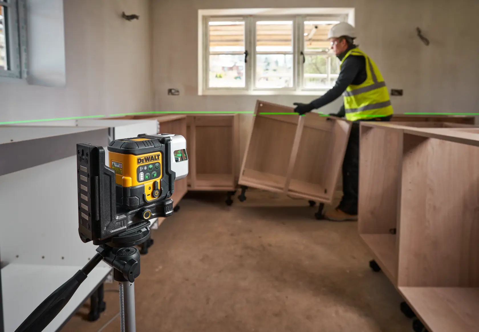 A DEWALT laser level device mounted on a tripod emits a green laser line across the room. In the background, a construction worker wearing a safety vest and helmet is installing wooden cabinets. The scene appears to be inside a partially completed room with a window and unfinished walls.
