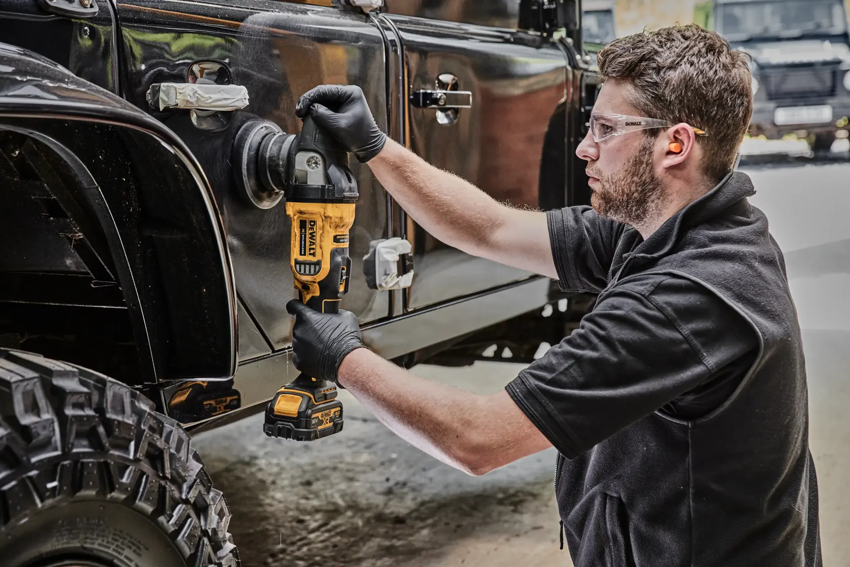 Una persona con guanti neri utilizza una lucidatrice a batteria DEWALT (modello DCM848N) per lucidare il lato di un veicolo fuoristrada nero. La scena si svolge in un'officina o garage.