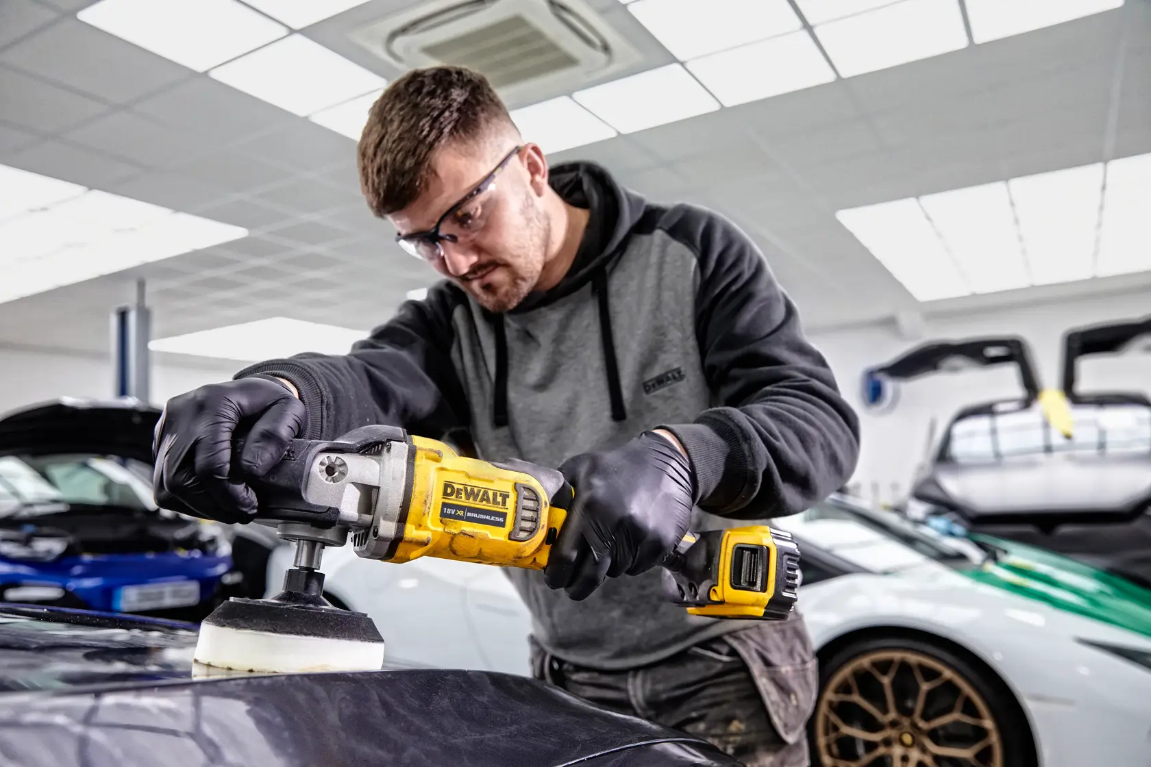 Persona usa lucidatrice DEWALT DCM849P2 su auto in officina moderna con veicoli di lusso sullo sfondo.