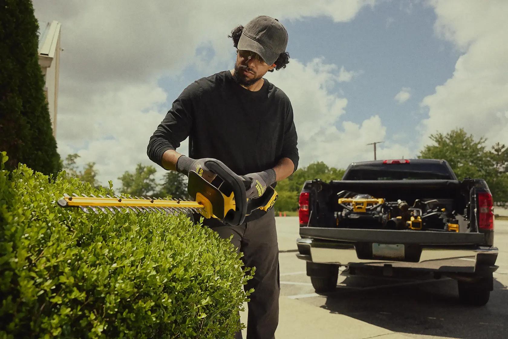 Persona pota siepe con tagliasiepi DEWALT a batteria, pickup con attrezzi sullo sfondo all’aperto.