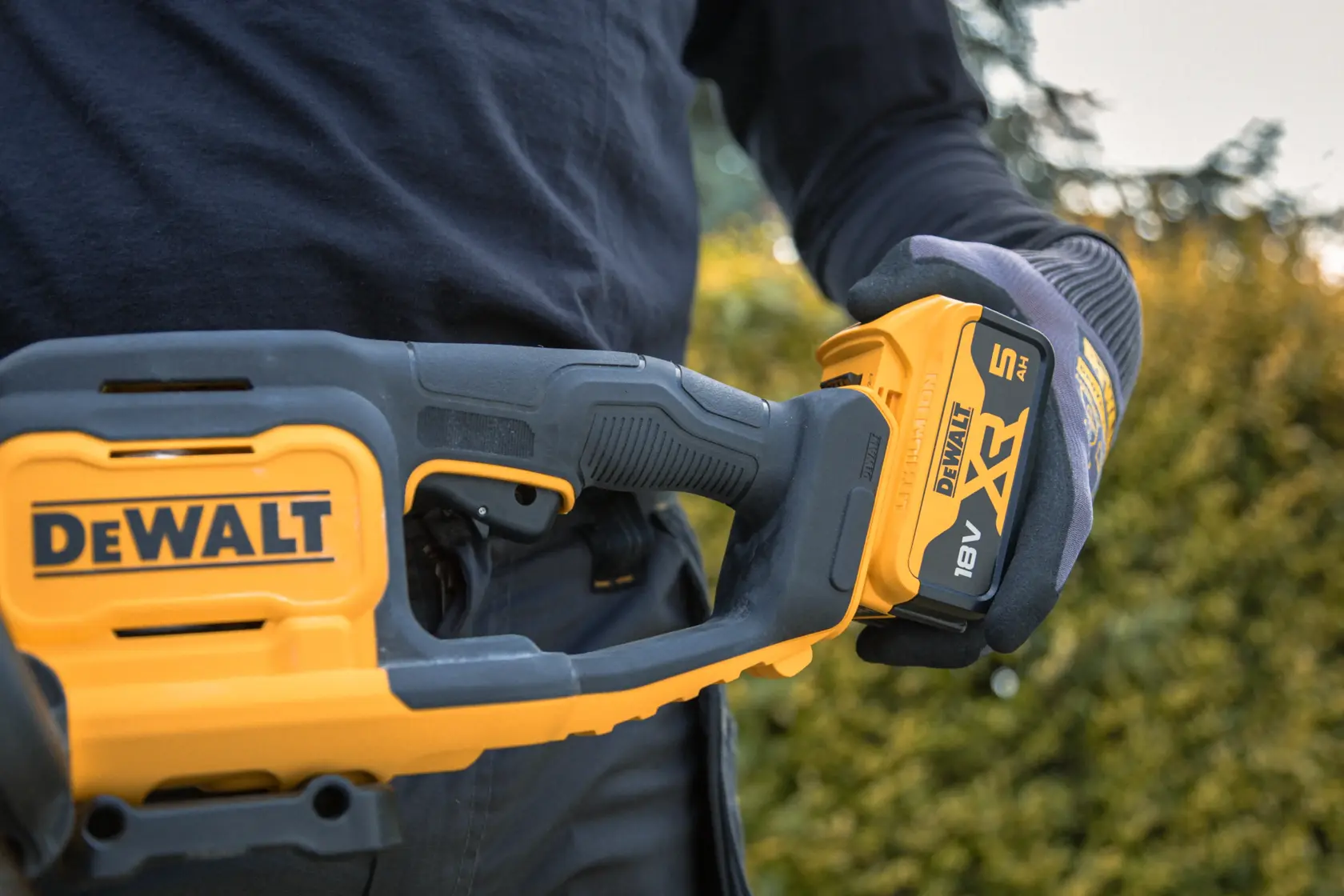 Close-up of a person holding a DEWALT cordless power tool outdoors, featuring a 18V XR 5Ah battery.