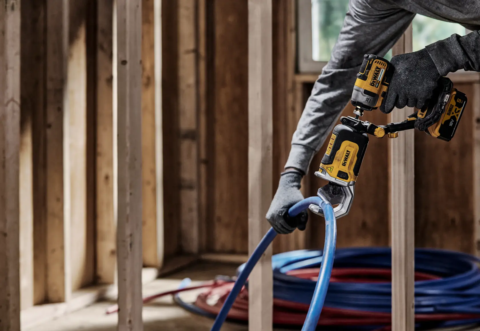 Una persona con i guanti utilizza un utensile elettrico DEWALT DT20560 per tagliare un tubo di plastica blu in una struttura interna in legno.