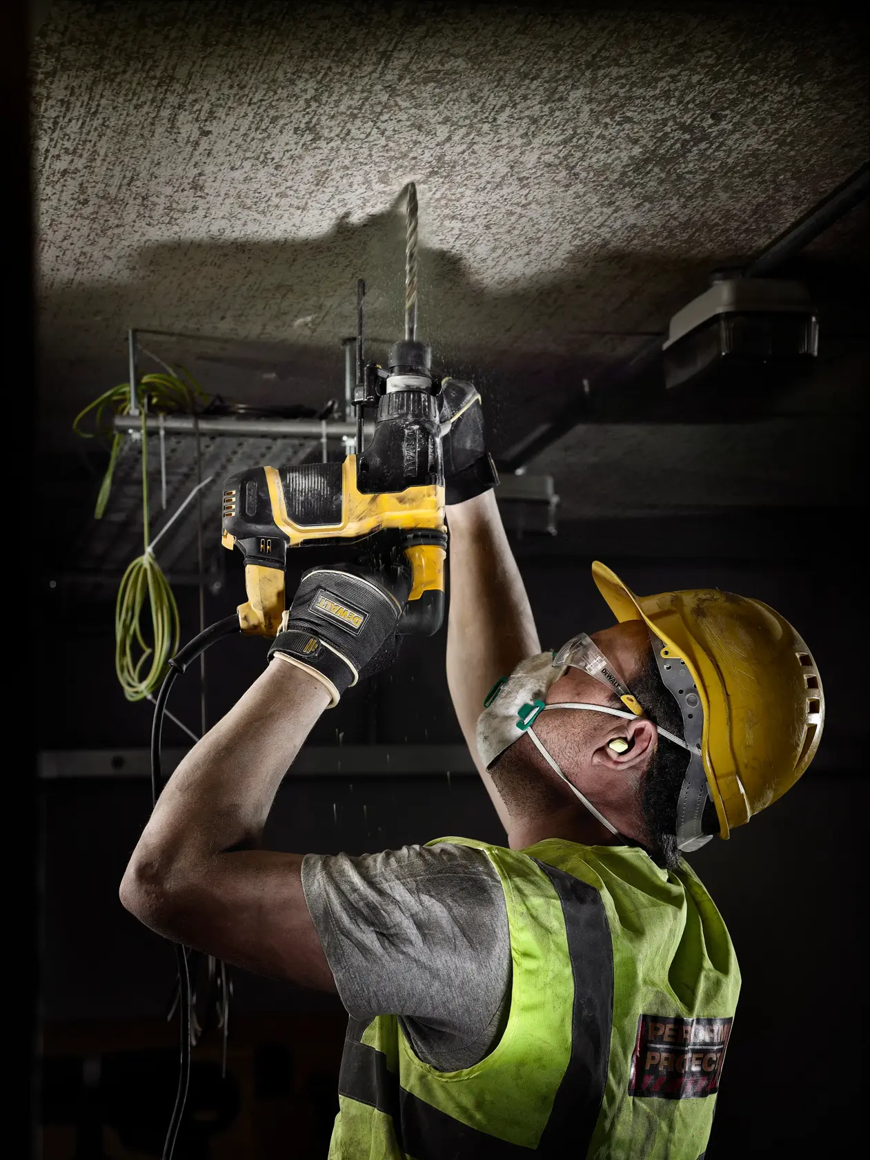 Worker in safety gear uses DEWALT electric drill to bore into ceiling on construction site.