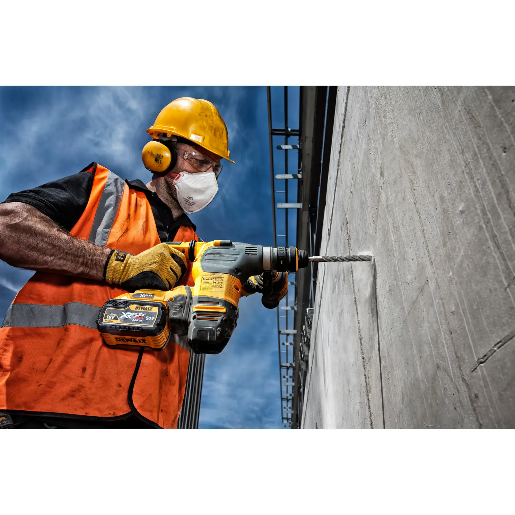 A construction worker wearing a yellow hard hat, orange safety vest, and gloves is using a DEWALT XR cordless rotary hammer drill with a DT9529 drill bit to drill into a concrete wall outdoors.