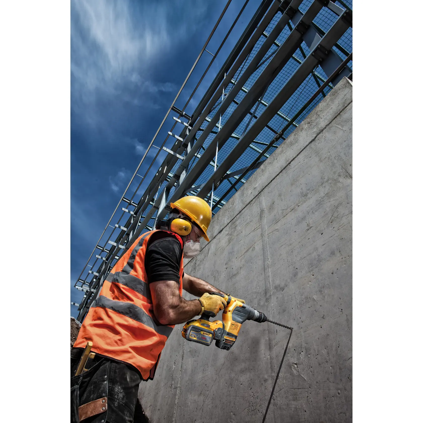 A construction worker wearing a yellow hard hat, ear protection, safety vest, and gloves is using a DEWALT power drill to work on a concrete wall at a construction site under a blue sky.