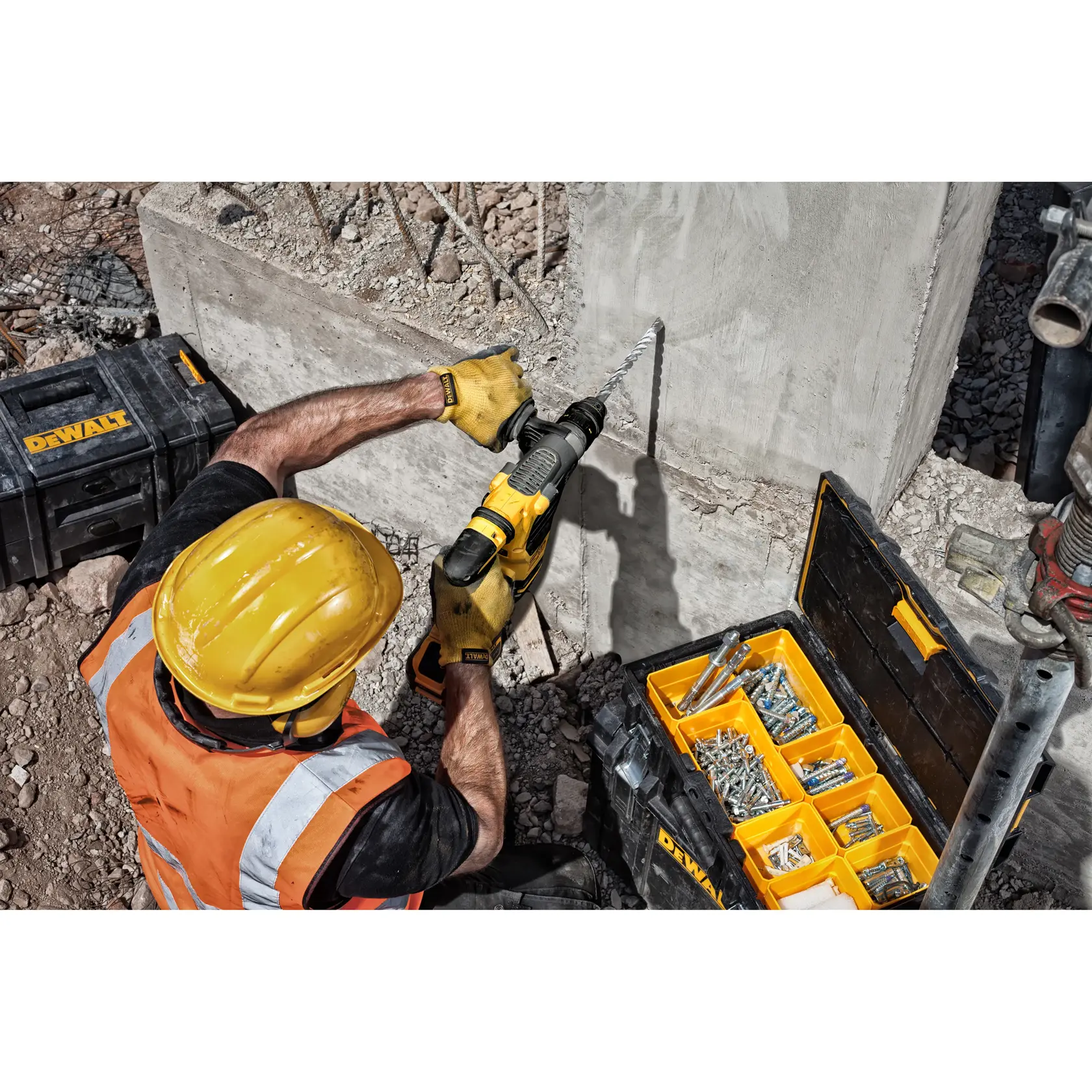 A construction worker wearing a yellow hard hat and orange safety vest uses a DEWALT power drill with a long drill bit to work on a concrete wall. Next to him are DEWALT toolboxes, one with organized compartments containing various screws and fasteners.