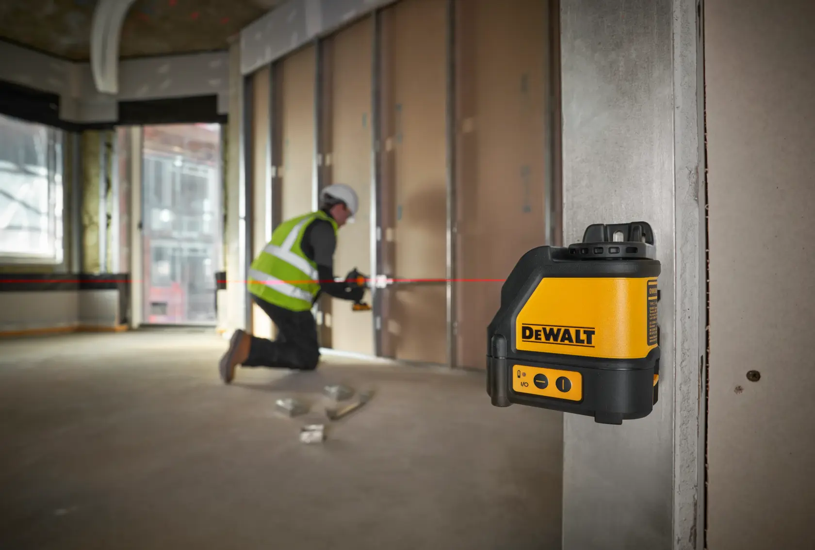 A DEWALT DW088K laser level is mounted on a metal column, projecting a red horizontal line across a construction site interior. In the background, a worker wearing a safety vest and helmet is aligning materials along the laser line.