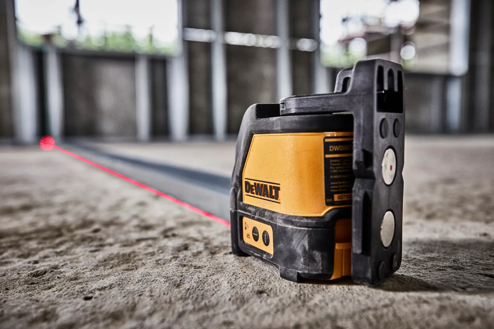 A DEWALT DW088K self-leveling cross line laser tool is shown projecting a red laser line on the floor inside an unfinished building with concrete walls.
