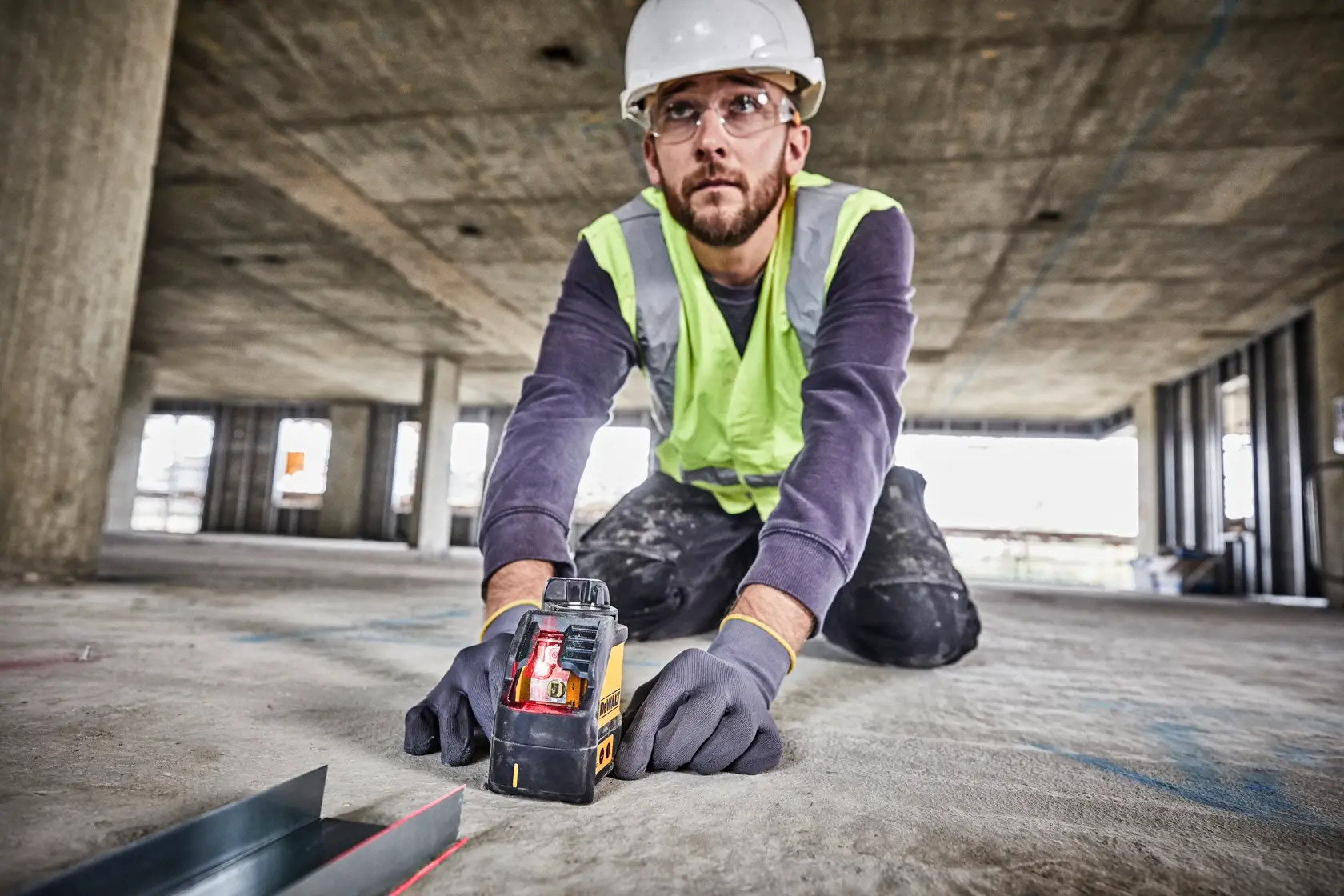 A person wearing a hard hat, safety vest, and gloves is kneeling on the floor of a construction site, using a DEWALT DW088K laser level. The device is projecting a visible red laser line for alignment. The surroundings show an unfinished building with concrete columns and open windows.