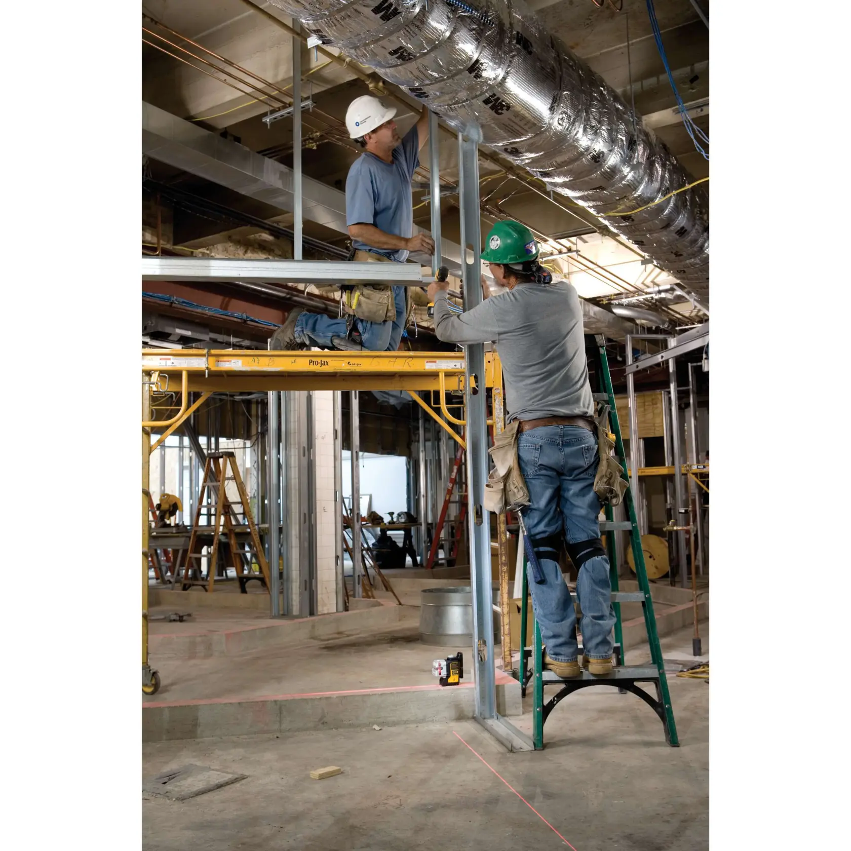 Deux ouvriers du bâtiment assemblent des structures métalliques sur un chantier intérieur. Un ouvrier est debout sur une échelle verte, tandis que l'autre est agenouillé sur un échafaudage jaune. Un niveau laser DEWALT DW089K est posé au sol, projetant des lignes laser rouges pour aider à l’alignement. L’espace de travail est rempli d’équipements de construction, de conduits et d’échafaudages.
