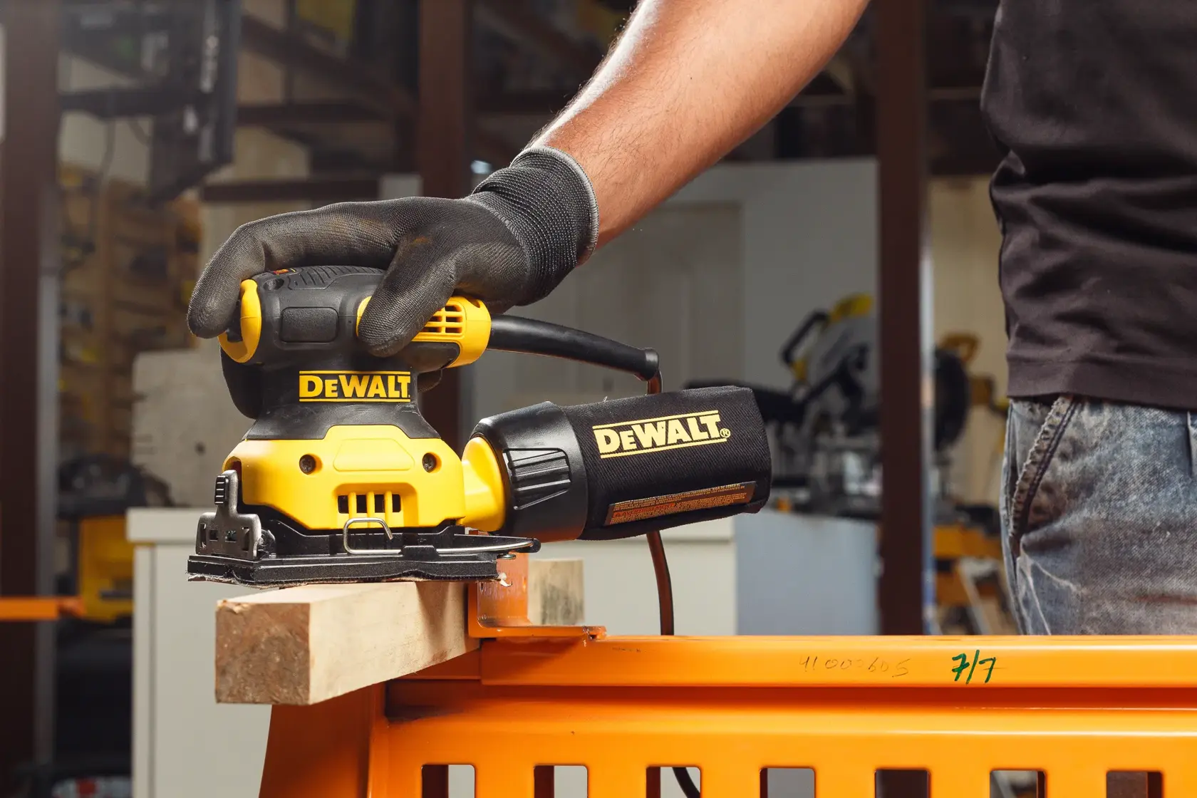 A close-up view of a gloved hand operating a yellow and black DEWALT DWE6411 electric sander on a wooden surface outdoors.
