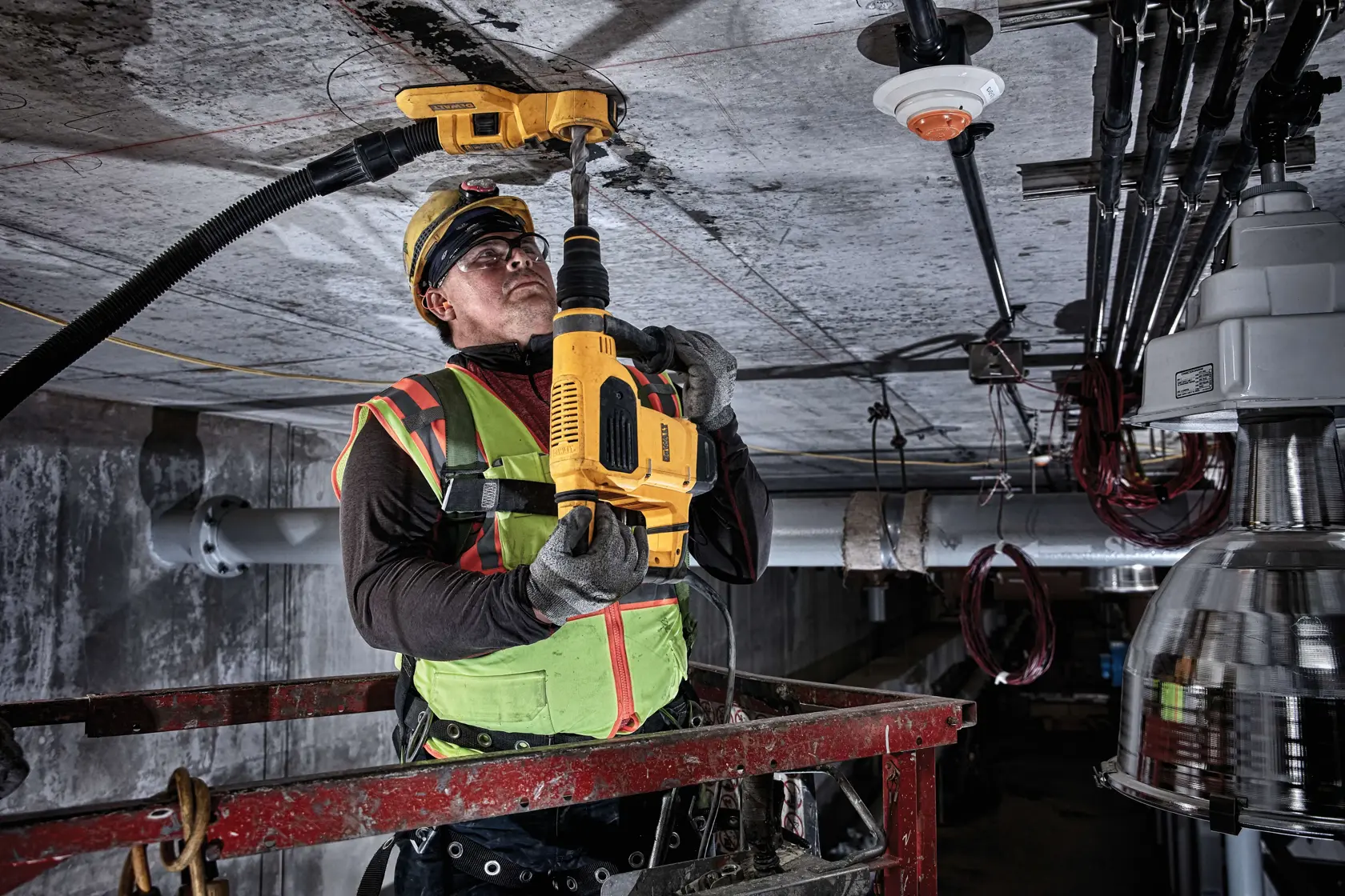 A construction worker in safety gear uses a DEWALT DWH050 tool for drilling into a ceiling in an industrial setting. The worker is standing on a lift platform, and a dust extraction attachment is connected to the tool.