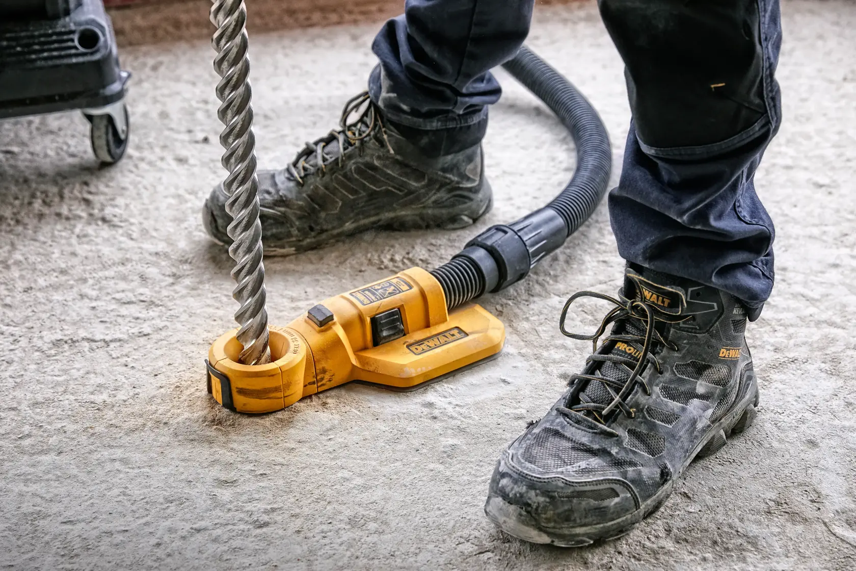 A DEWALT DWH050 dust extraction attachment is connected to a vacuum hose and positioned around a large drill bit on a construction site floor. The feet of a person wearing DEWALT branded work boots and dark pants are visible.