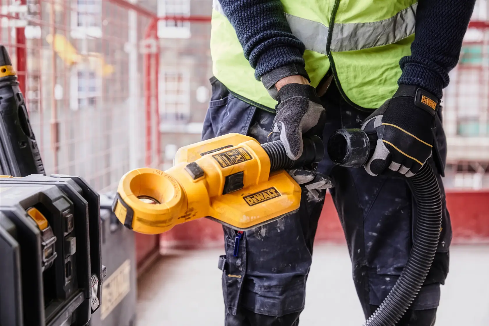 A person wearing a safety vest and gloves is using a DEWALT DWH050 dust extractor attachment at a construction site. The yellow tool is connected to a hose and positioned near a DEWALT storage case.
