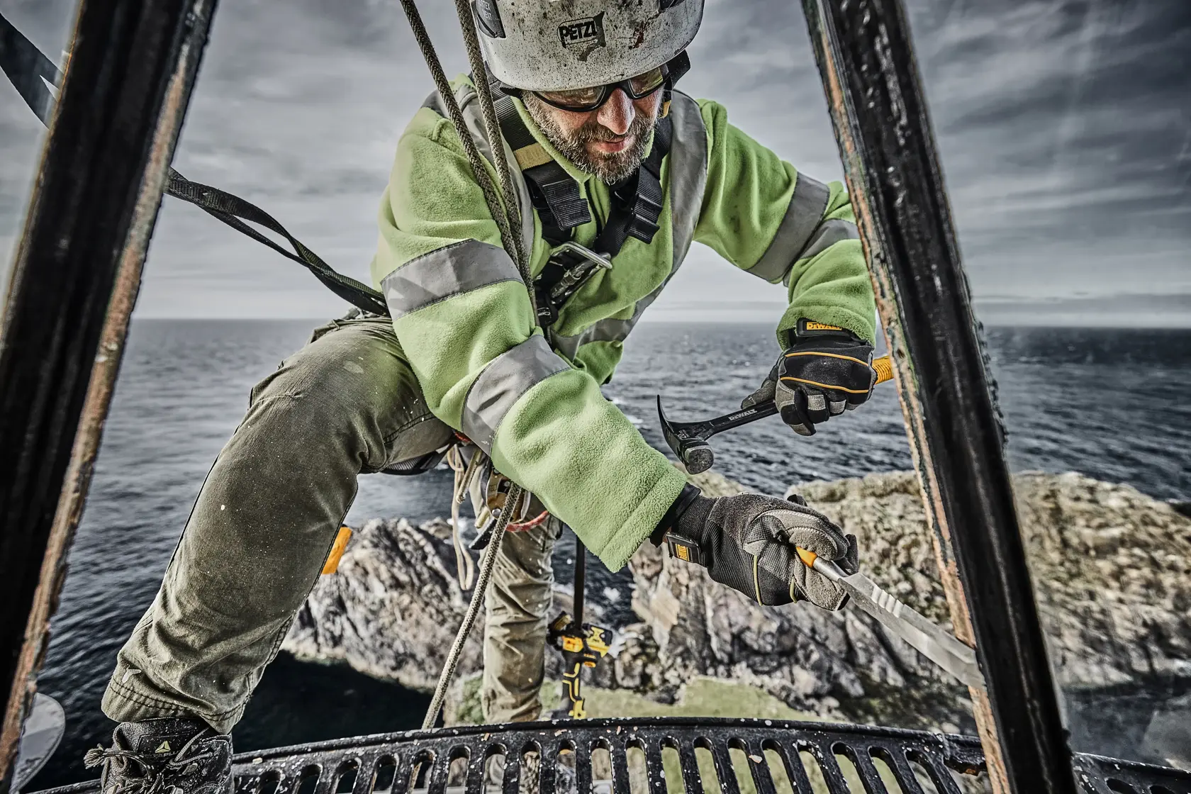 Operaio usa martello DEWALT su struttura metallica vicino al mare, con giacca verde e dispositivi di sicurezza.