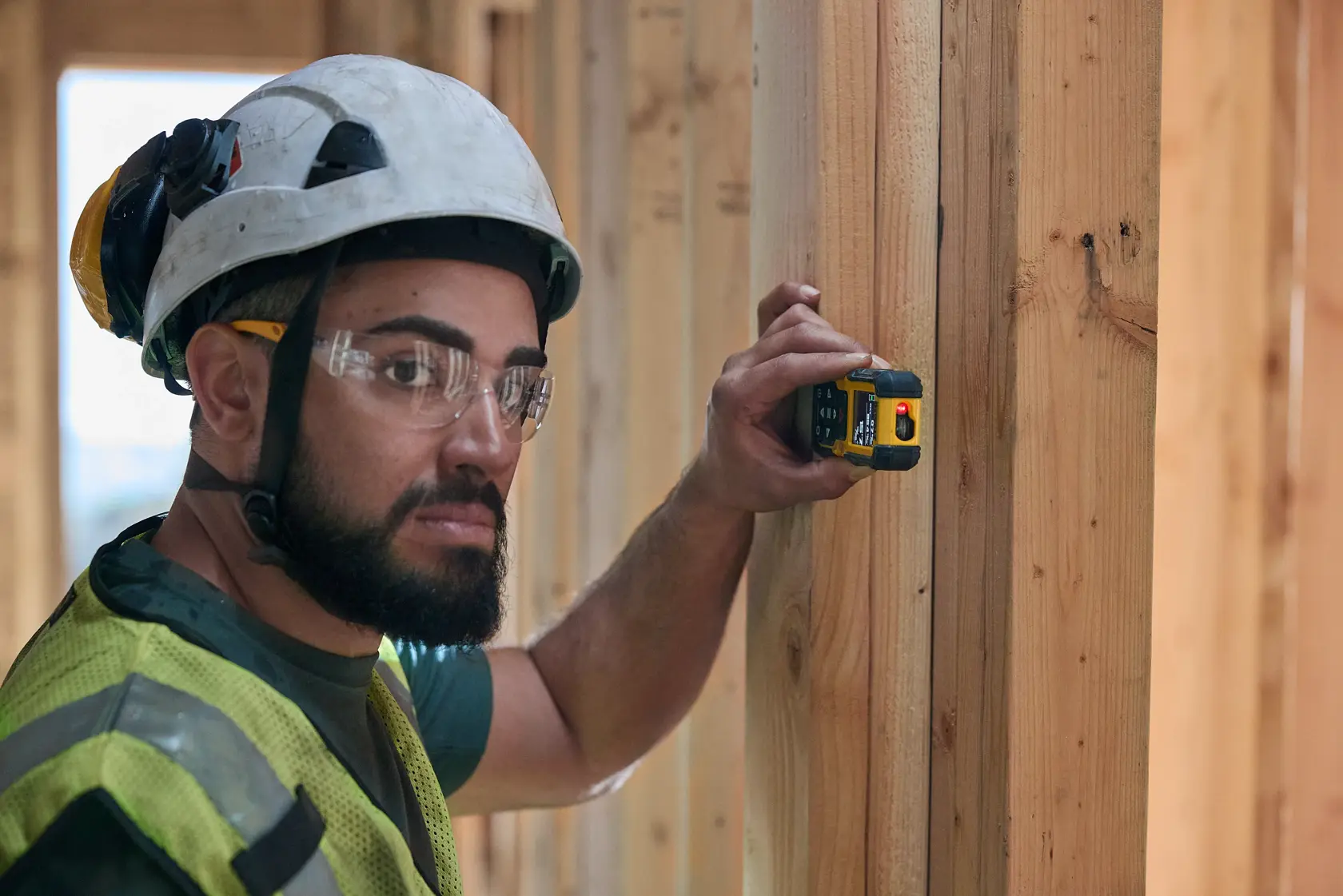 A construction worker wearing a safety helmet and reflective vest is using a DEWALT DWHT77200 laser distance measurer against a wooden beam inside a building under construction. The worker's face is obscured.