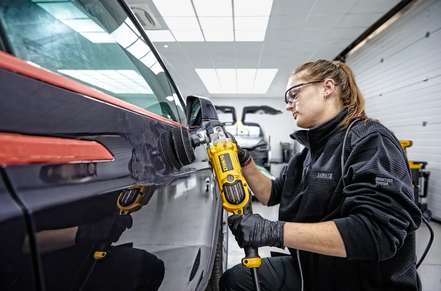 A person is polishing the hood of a red sports car with a DEWALT DWP849X polisher in a garage.