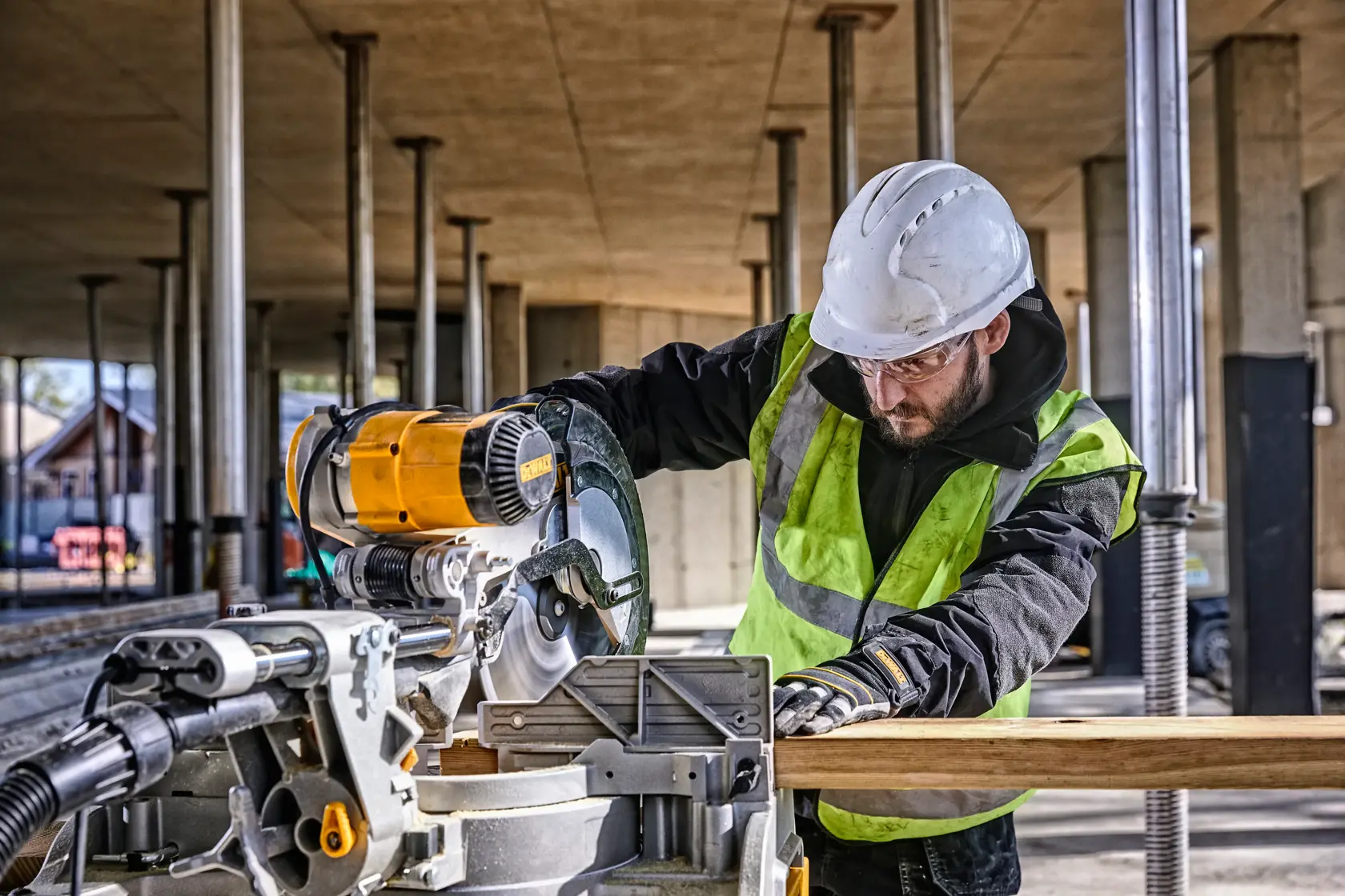 A construction worker wearing a helmet and high-visibility vest operates a DEWALT DWS780 miter saw on a building site. The saw is cutting a piece of wood, and there are metal columns and construction materials in the background.