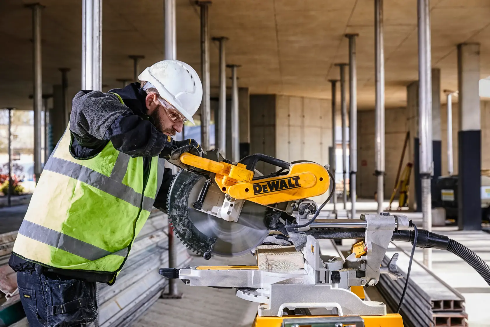 A DEWALT DWS780 miter saw being used to cut a wooden plank at a construction site. The operator is wearing a safety helmet, gloves, and a high-visibility vest. The background shows a partially constructed building with vertical pillars.